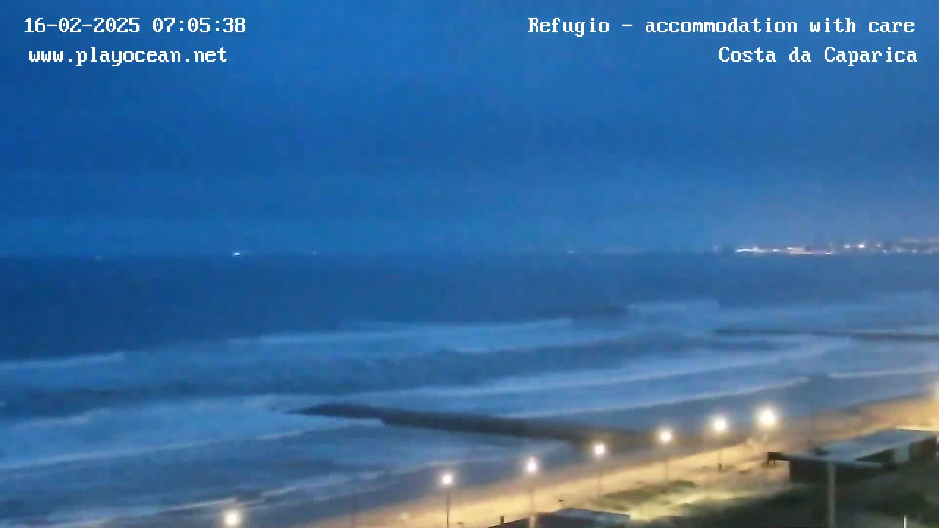 A dark-blue ocean with waves rolls onto a beach at dusk, illuminated by streetlights along a paved walkway.