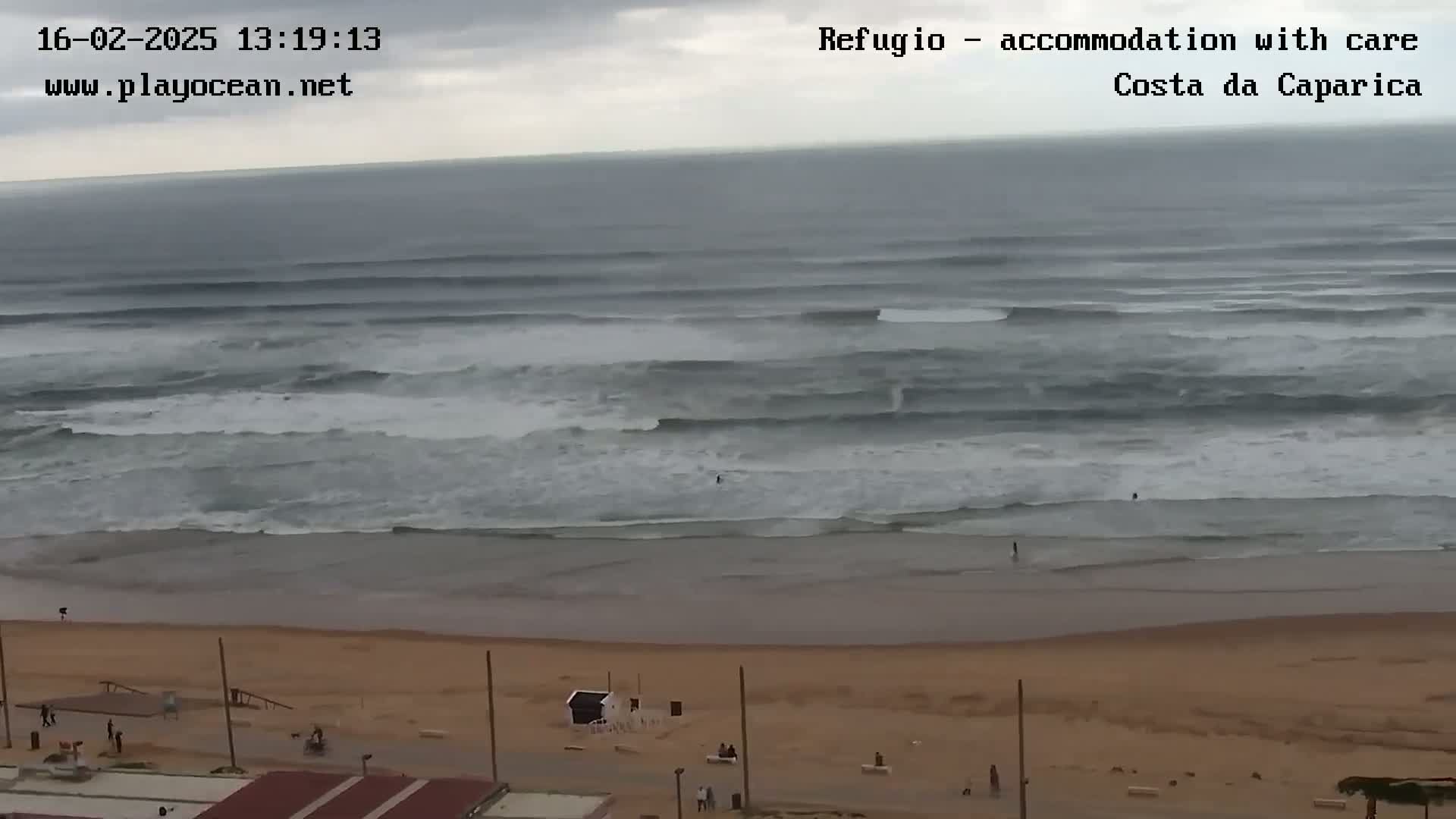 A wide shot shows a beach with moderate waves under an overcast sky, and a few people are visible on the sand.