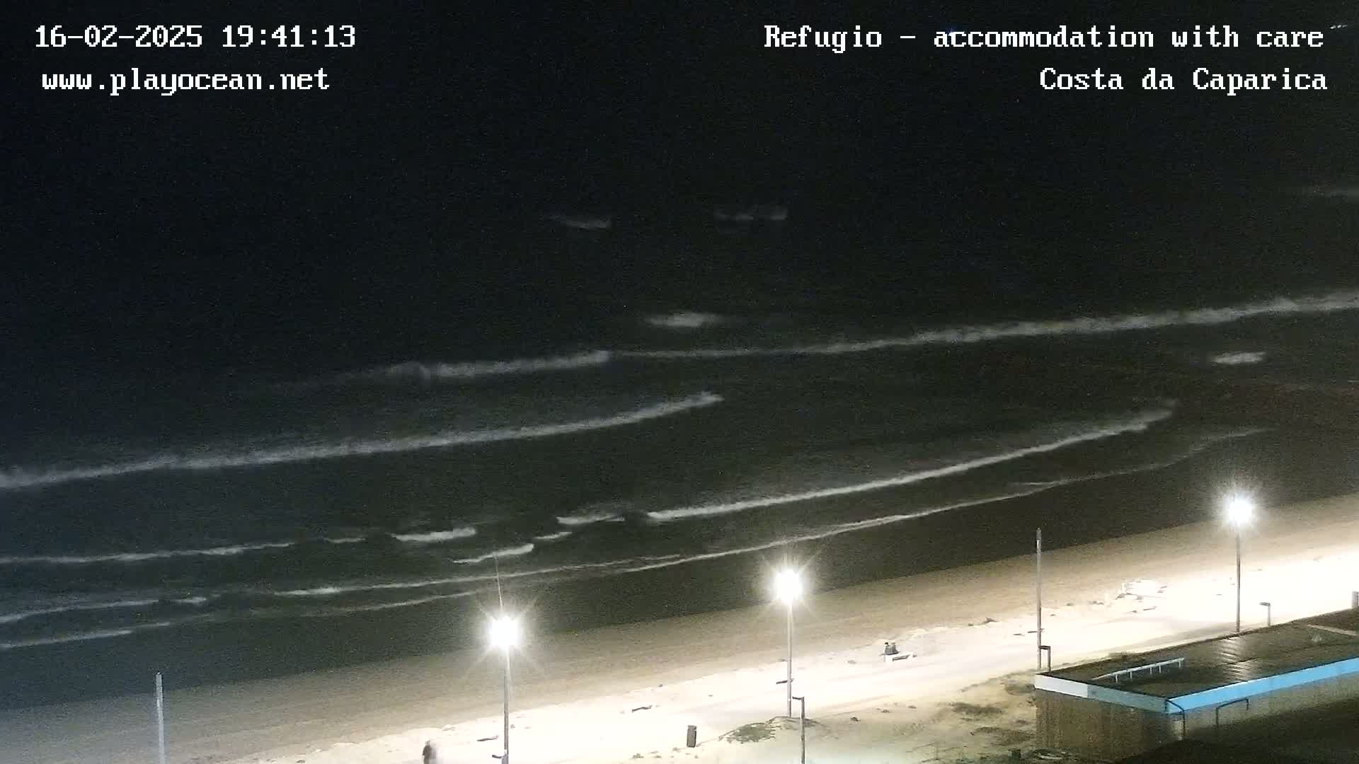 At night, moderate waves roll onto a sandy beach illuminated by streetlights.