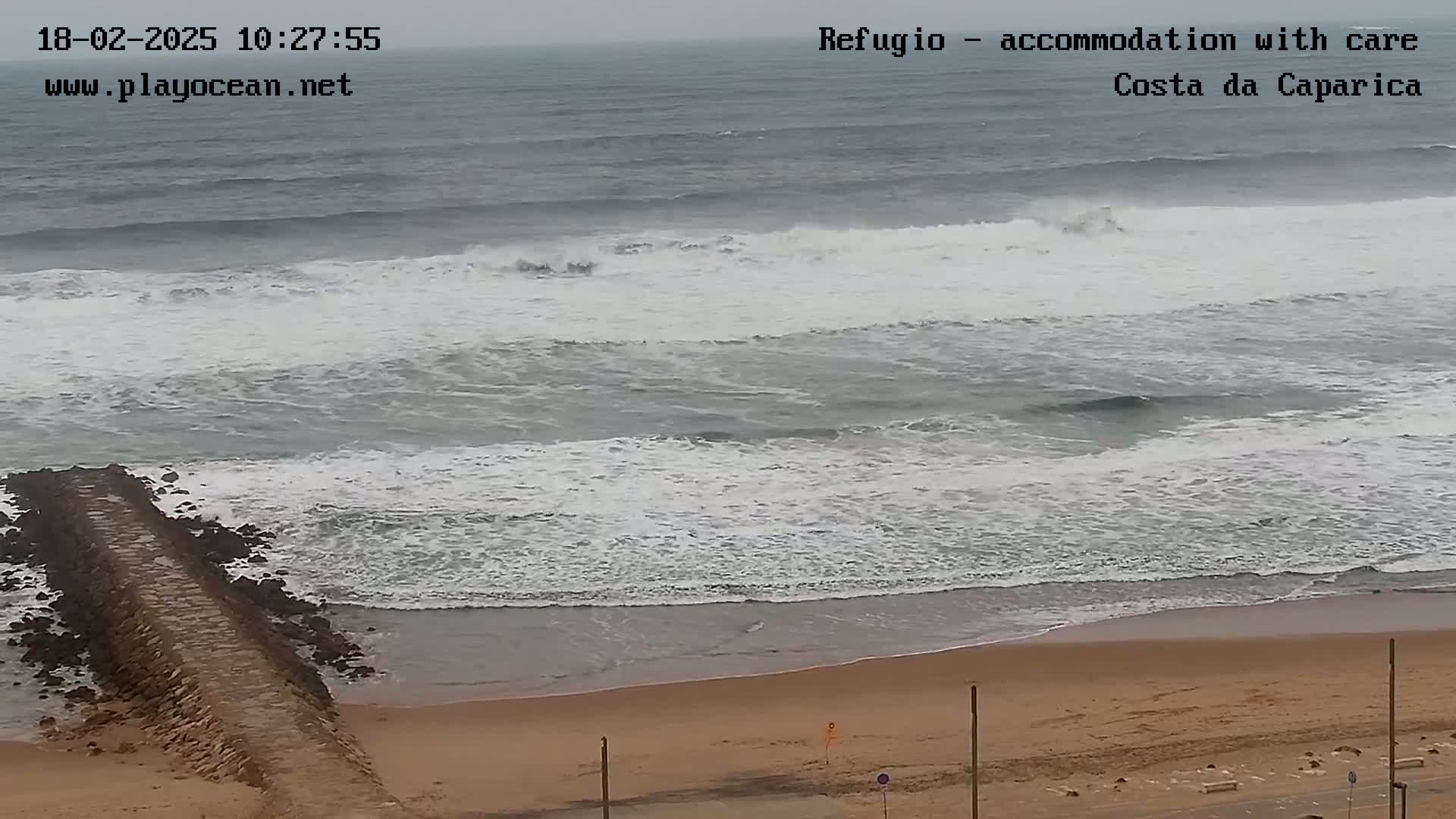 A long, low stone jetty extends into a gray, overcast ocean with moderate waves breaking on a sandy beach.