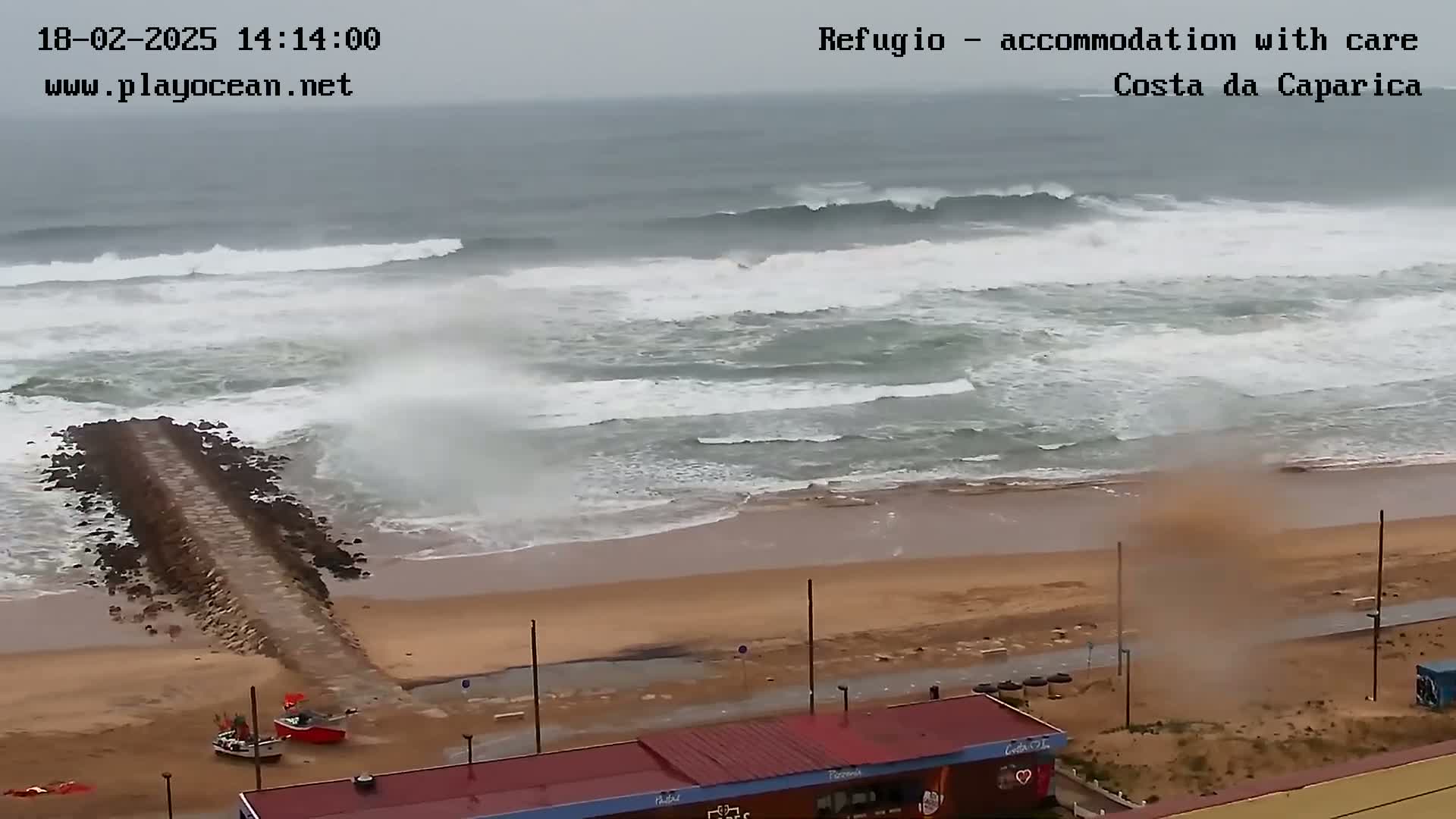 A sandy beach with a rocky jetty is seen under an overcast sky, with large waves crashing onto the shore and a small dust devil forming on the beach.