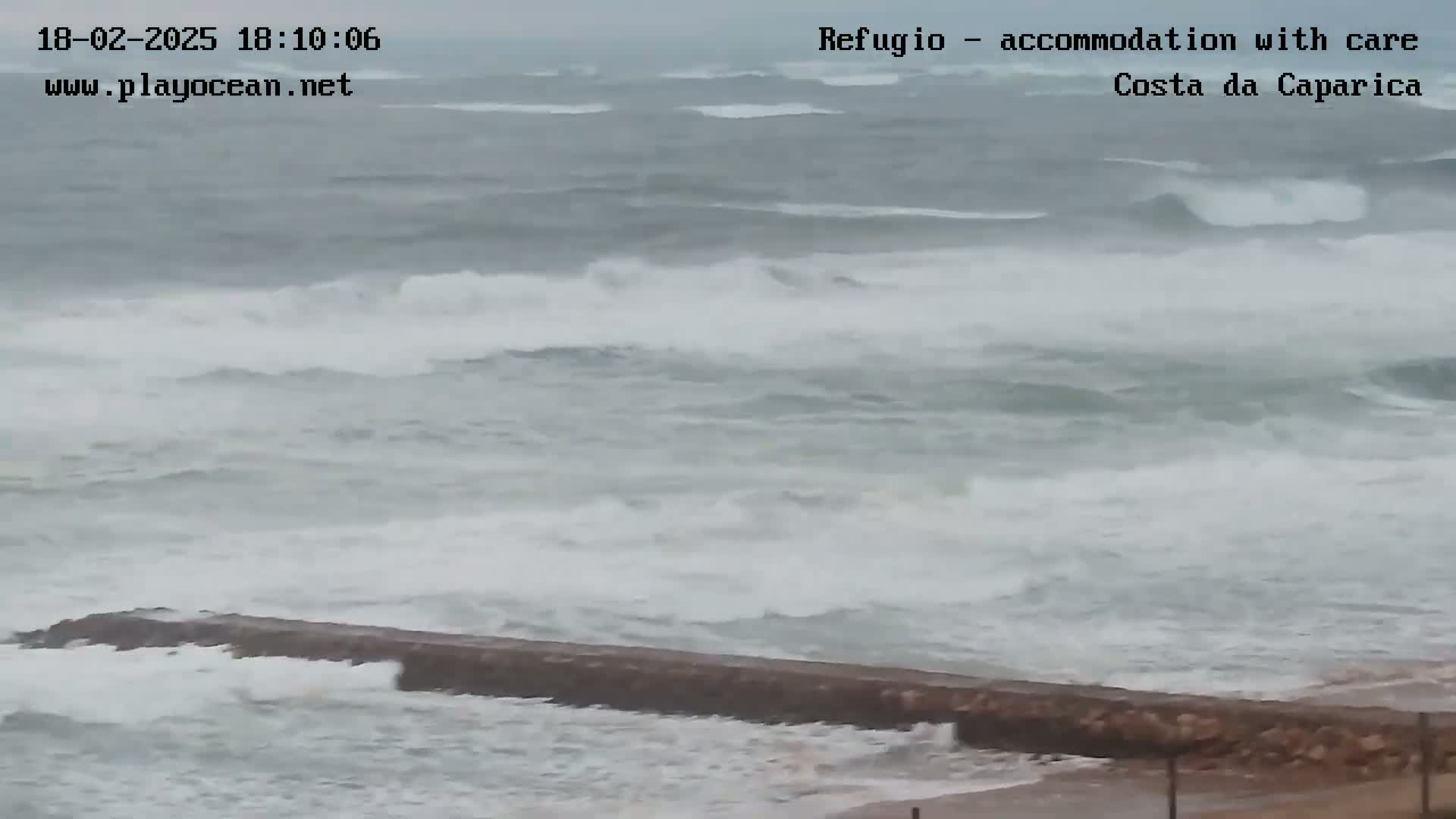 Rough ocean waves crash against a low-lying seawall during stormy weather.