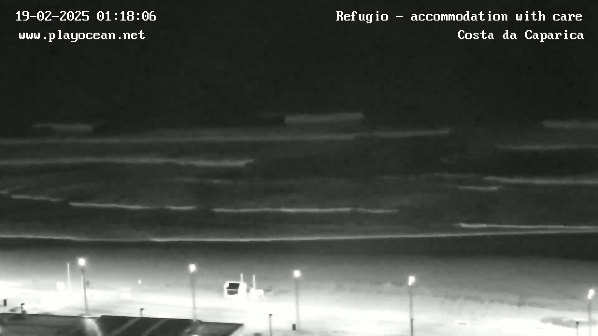 A nighttime, high-angle view shows a dark beach with gentle waves under a cloudy sky.