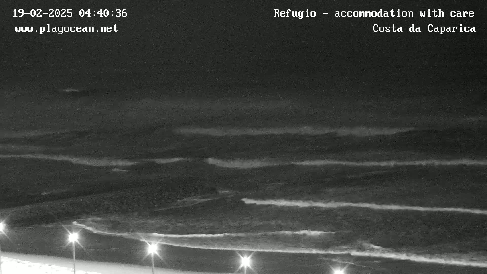 Under a dark night sky, gentle waves roll onto a beach illuminated by several lights.