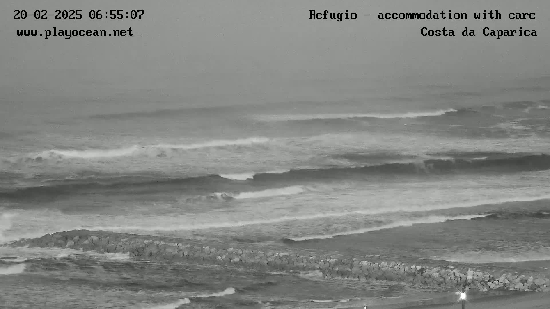 A grayscale image shows a rocky breakwater separating a beach from moderately sized waves in overcast conditions.