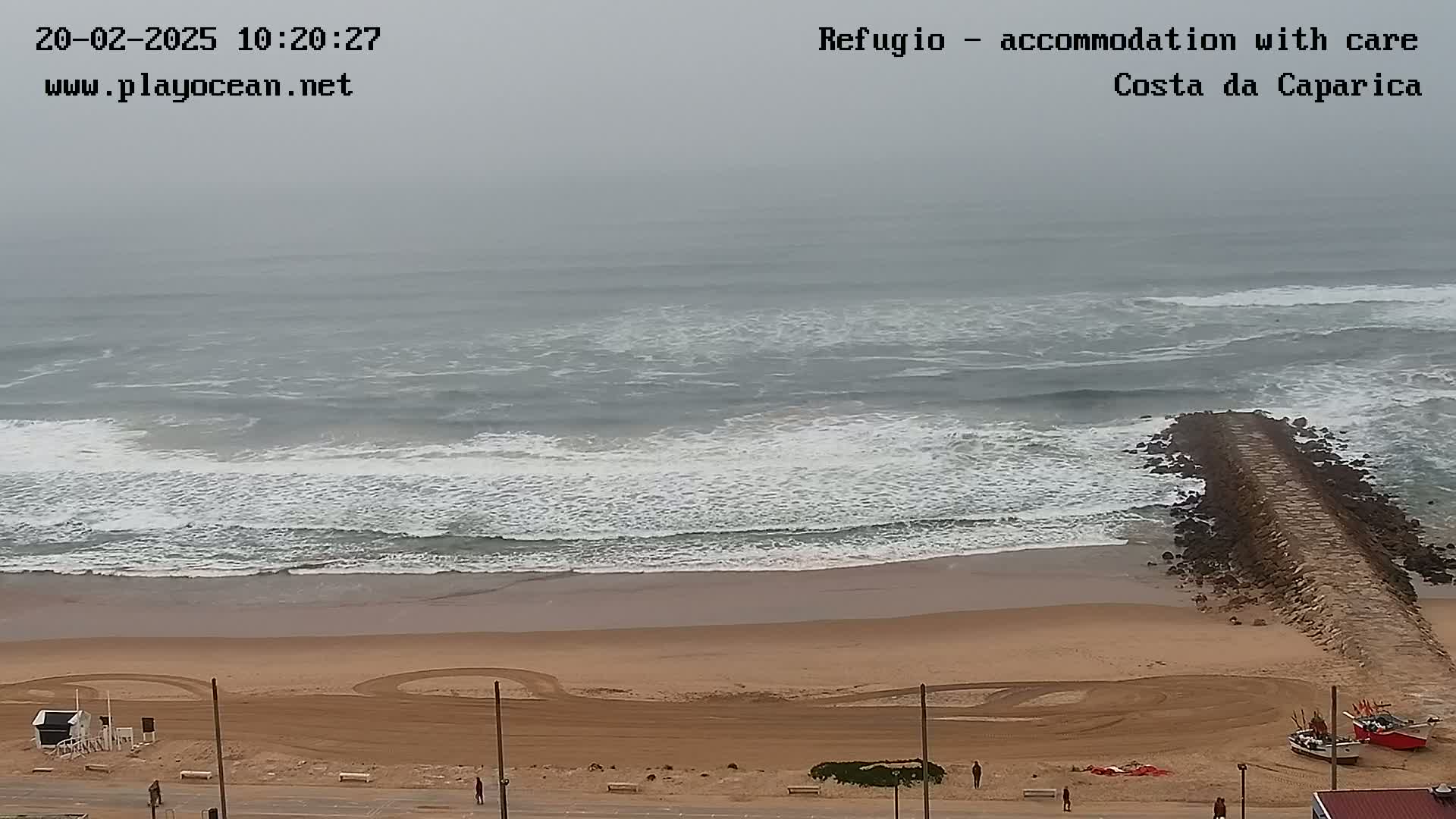 A long jetty extends from a sandy beach into a gray, overcast ocean with moderate waves.