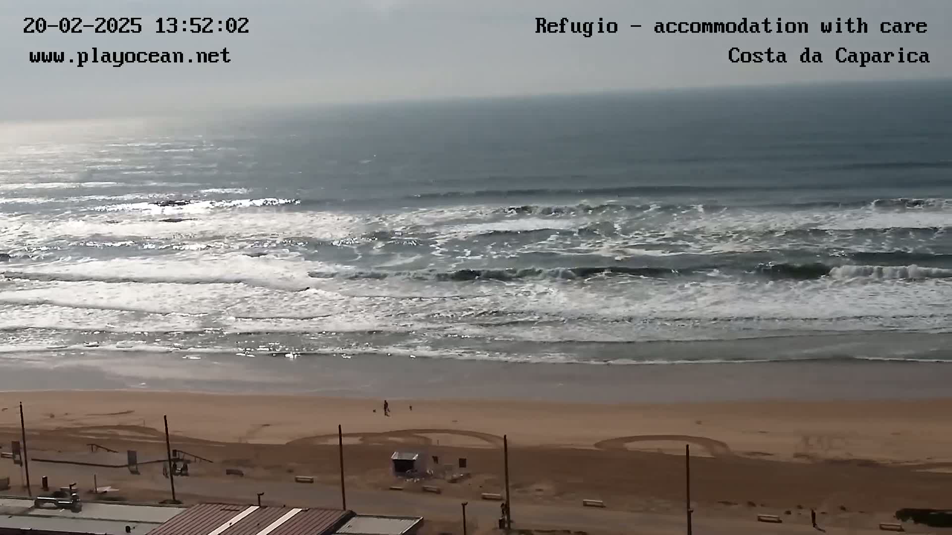 A mostly cloudy day shows moderate waves crashing onto a sandy beach with a few people visible near the water's edge and buildings in the foreground.