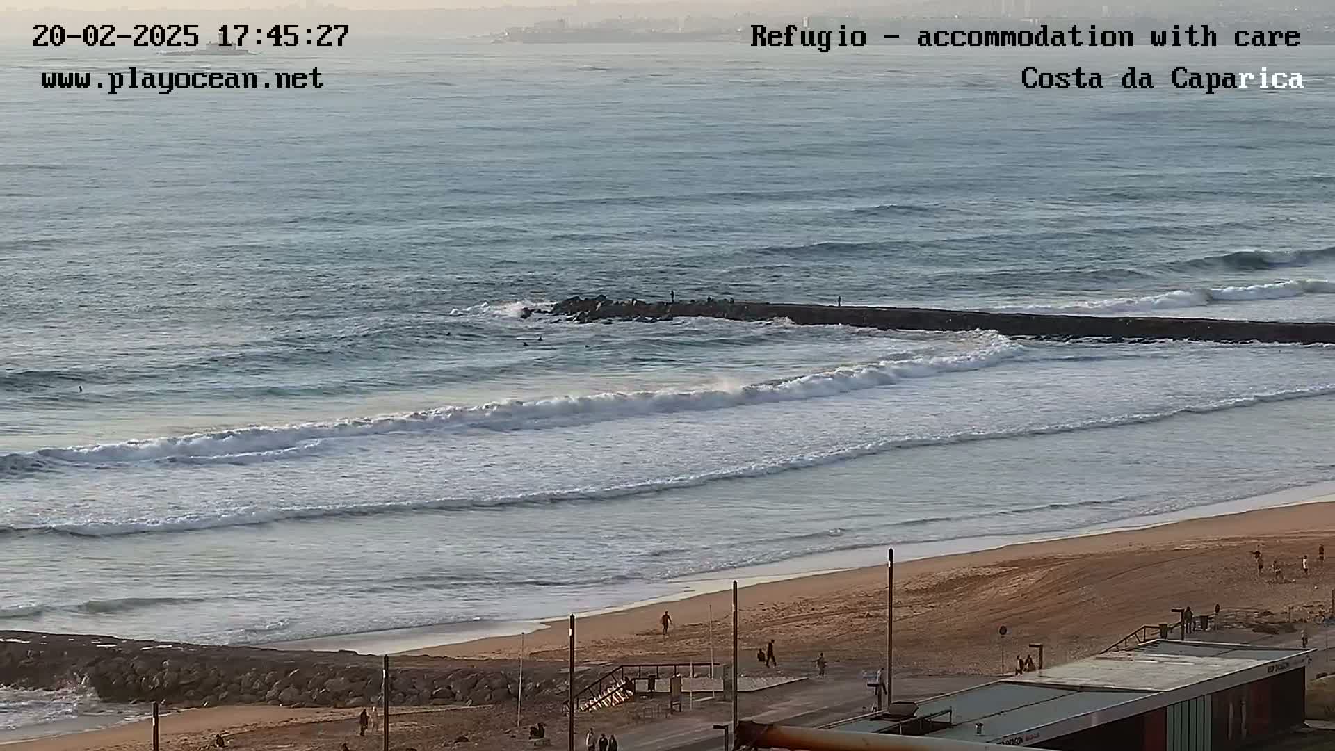 A long sandy beach with moderate waves rolling in under a partly cloudy sky, featuring a rock jetty and a few people on the beach and in the water.