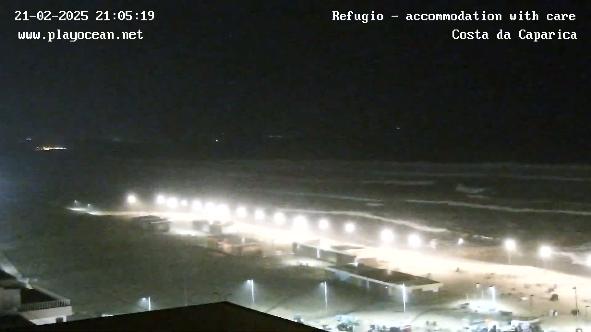 A nighttime view of a beach with moderately rough waves under hazy conditions, illuminated by lights along a boardwalk.