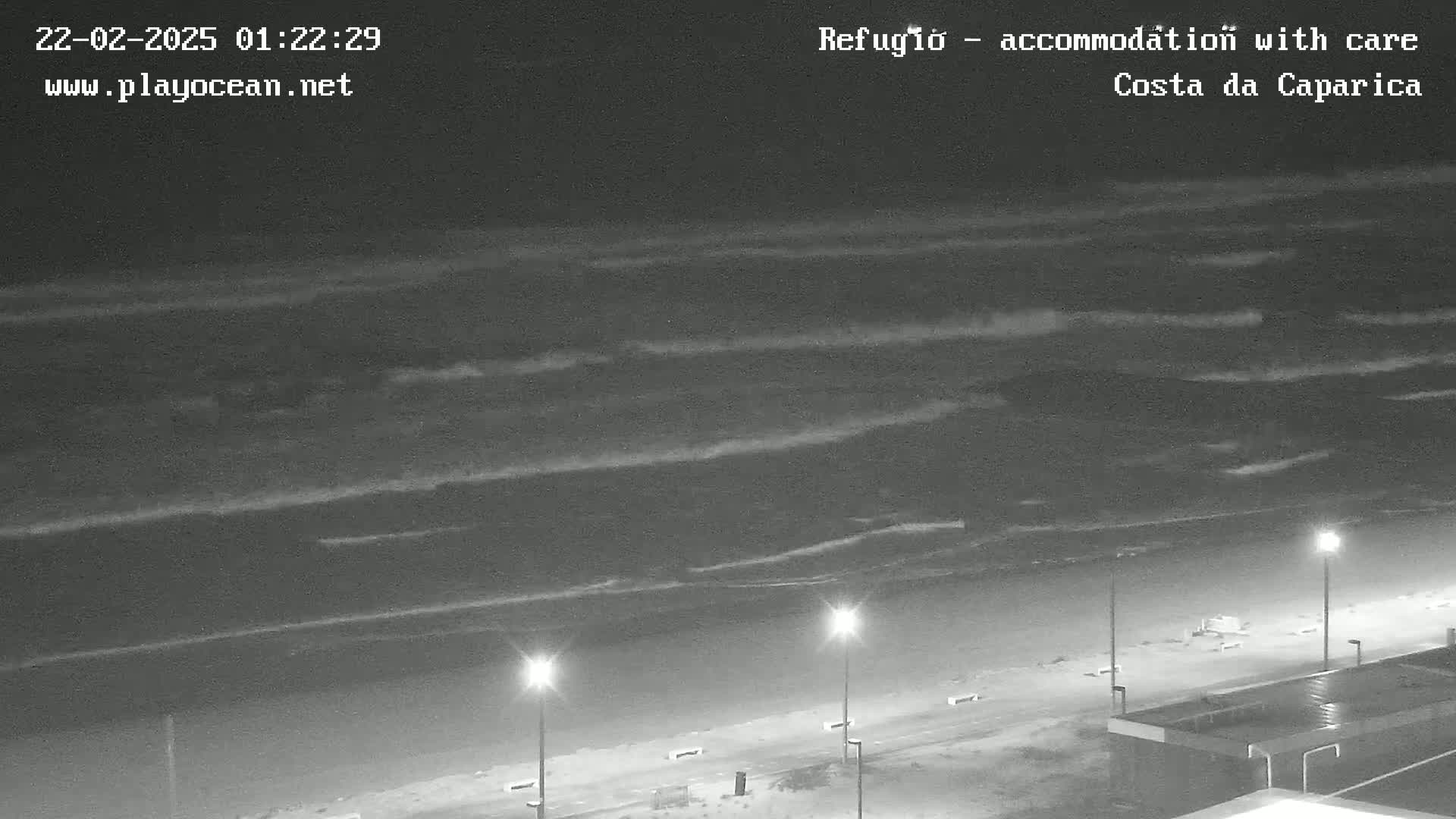 A nighttime view shows dark waves rolling onto a beach with several lights illuminating a mostly empty boardwalk.
