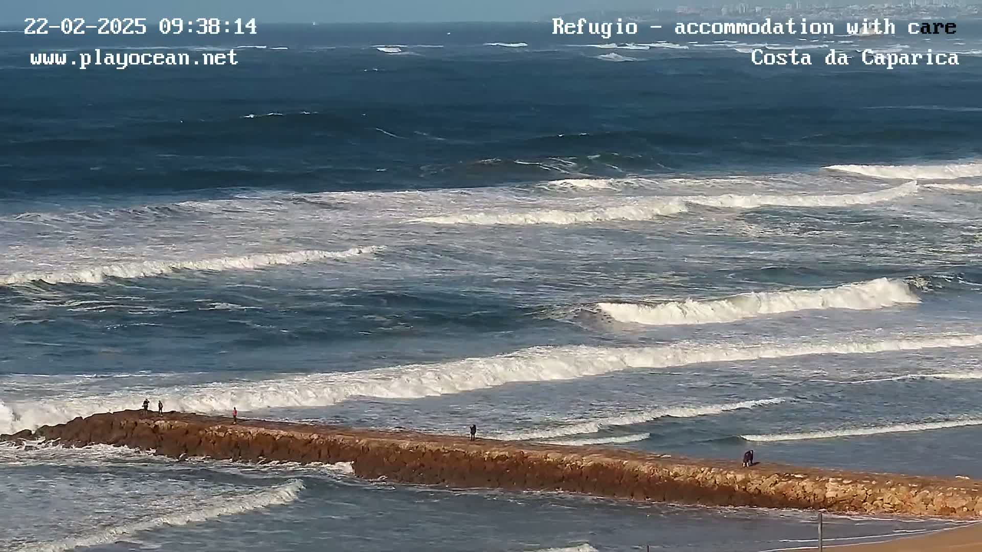 A long, low rock jetty extends into a wavy ocean under a clear sky.