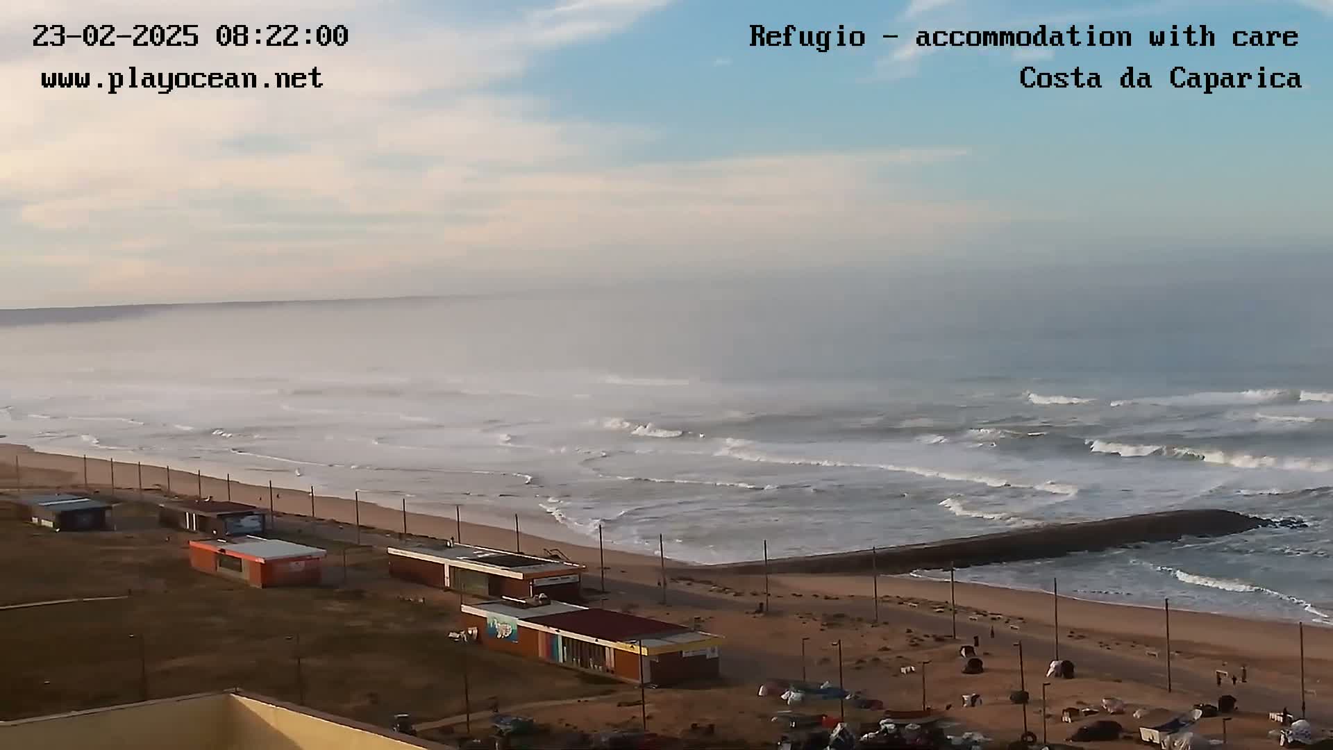 A foggy morning shows a beach with moderate waves crashing near a breakwater, several small buildings along the shore, and a mostly empty parking lot.