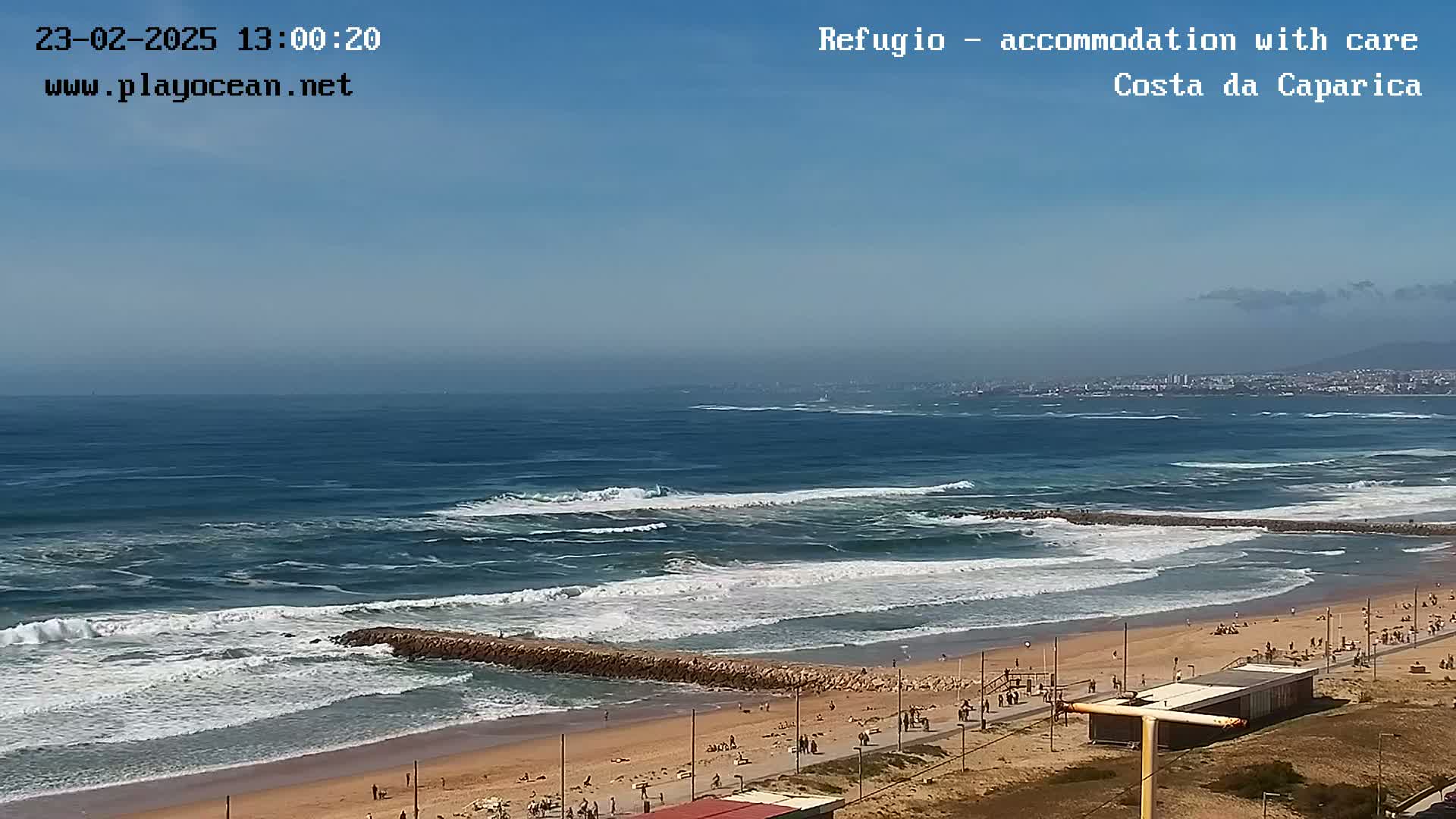 A sandy beach with a rocky breakwater extends into a blue ocean with moderate waves under a clear sky.