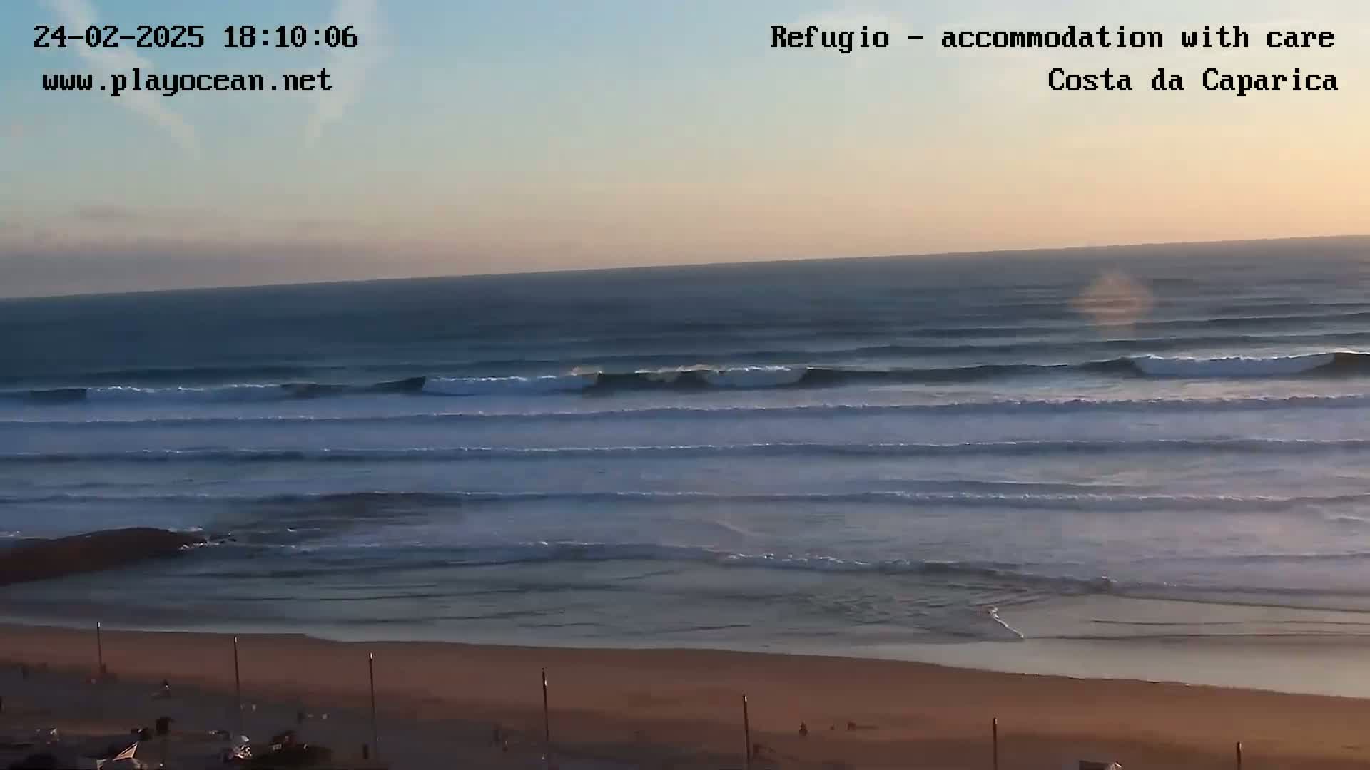 A sandy beach with a calm ocean under a clear, sunset sky shows several consistent waves rolling into shore.