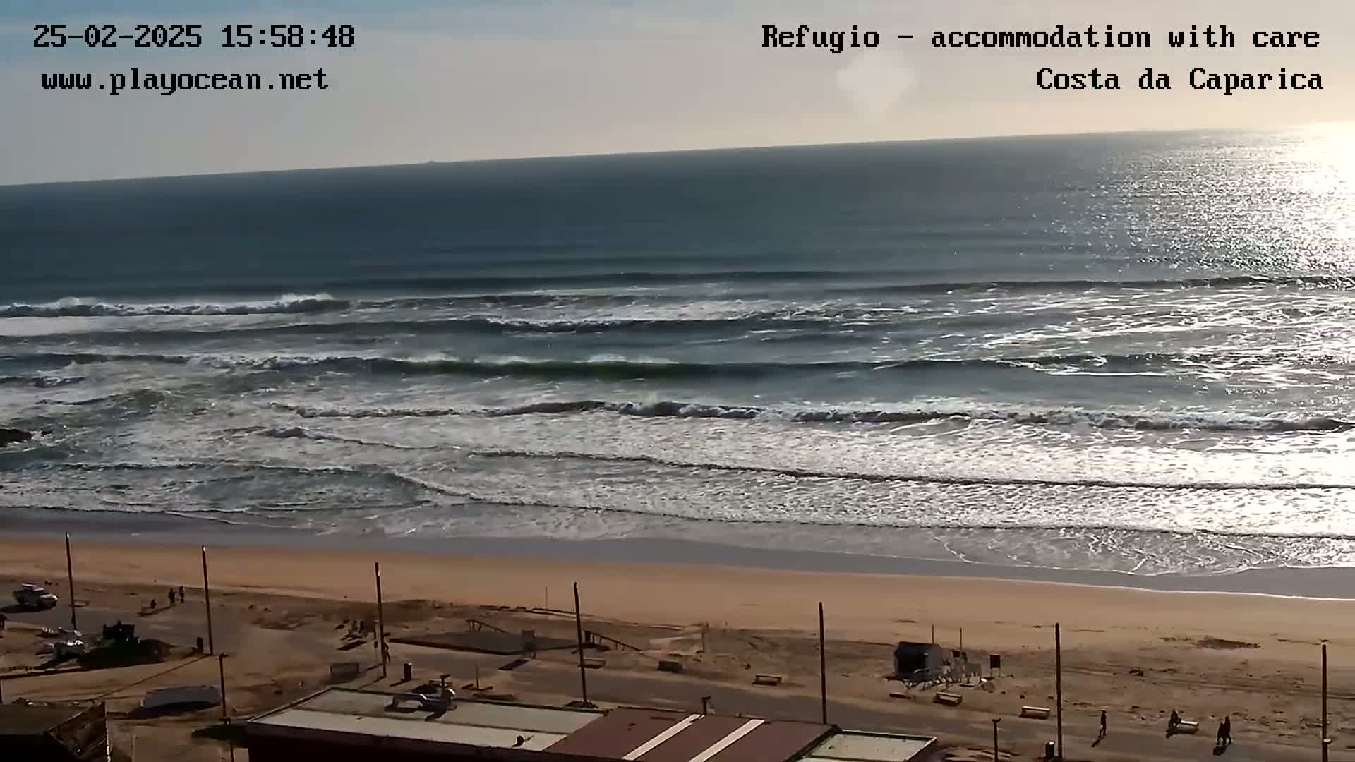 A long, sandy beach with moderate waves under a mostly clear sky is visible from a high vantage point, showing a coastal town in the foreground.