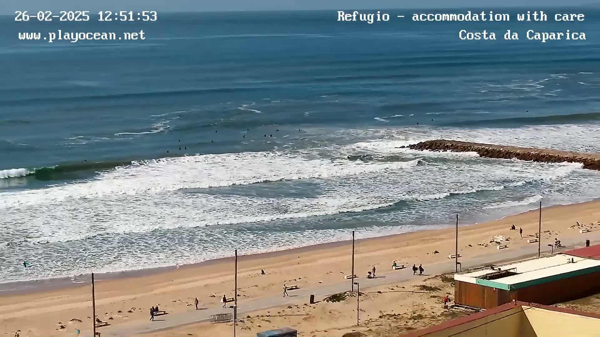 A sandy beach with a paved walkway and several people, is next to a blue ocean with waves breaking near a low-lying rock jetty, under a sunny sky.