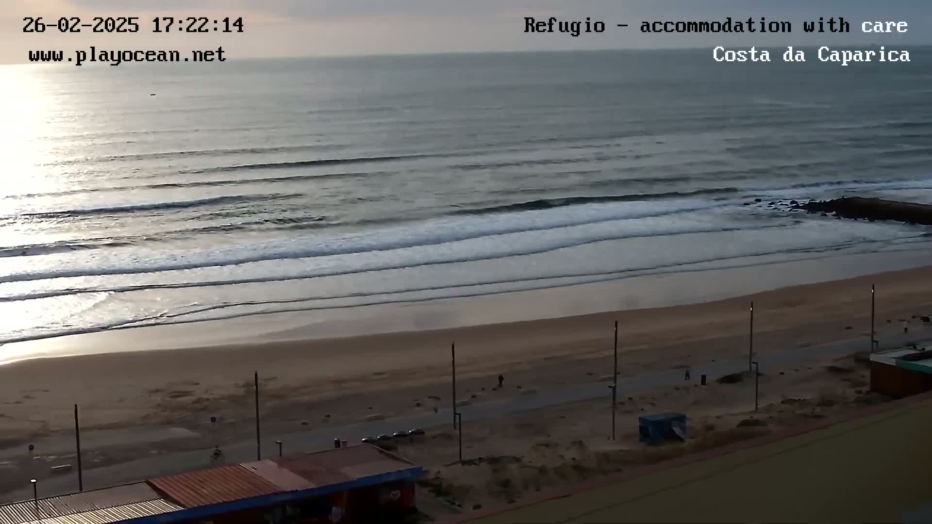 An overcast sky overlooks a sandy beach with calm waves rolling onto the shore, and a few people are visible walking along the beach.
