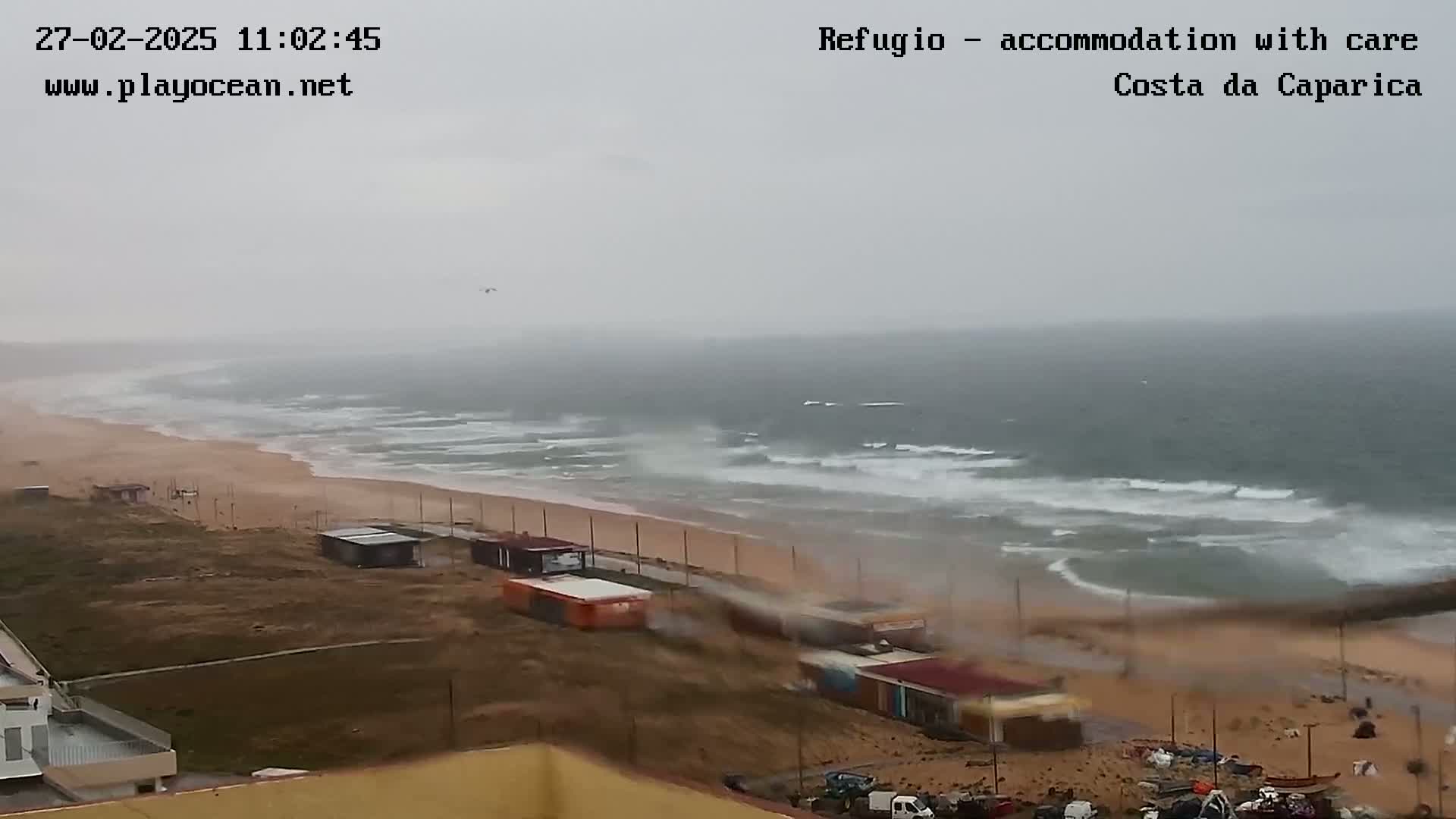 A beach scene shows a grey, overcast sky and choppy ocean waves crashing on a sandy shore near buildings.