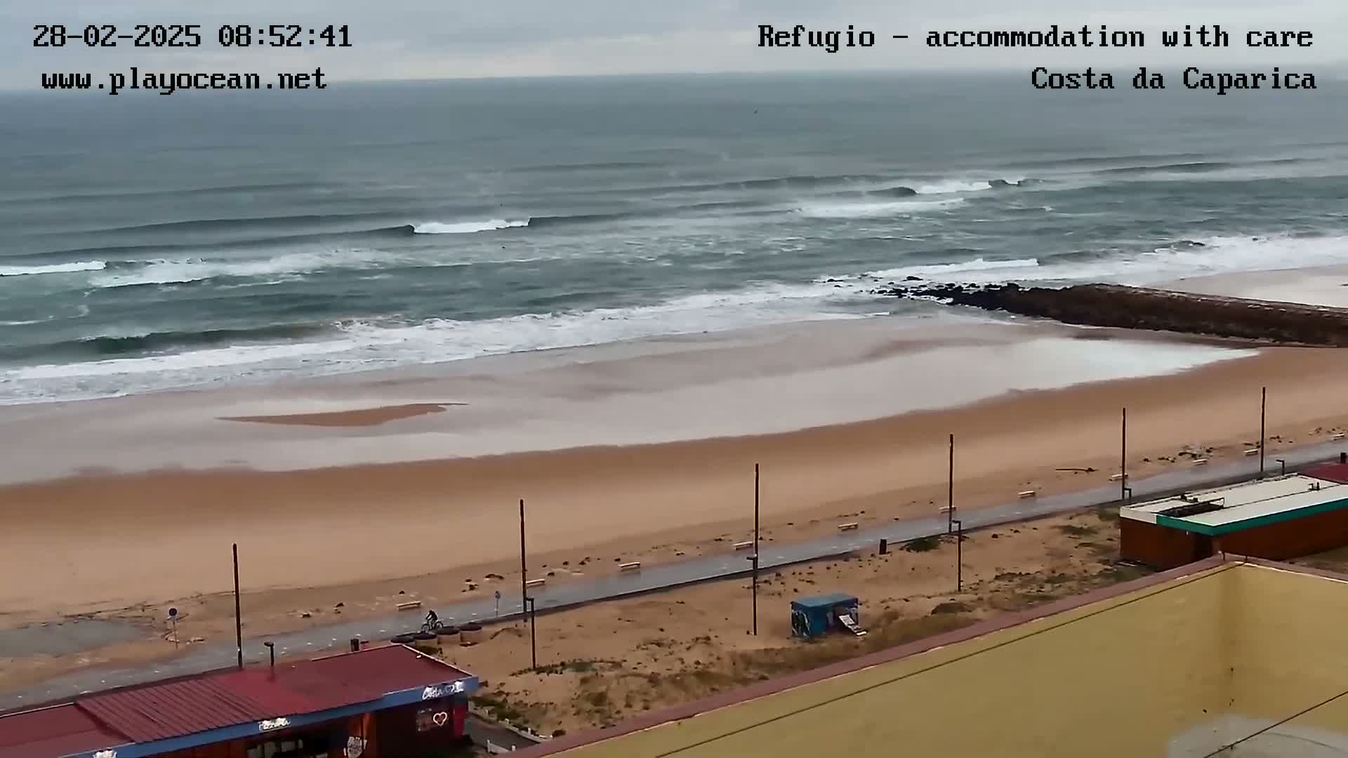 A sandy beach with a paved walkway and buildings is shown, with an overcast sky and moderate waves rolling into the shore.