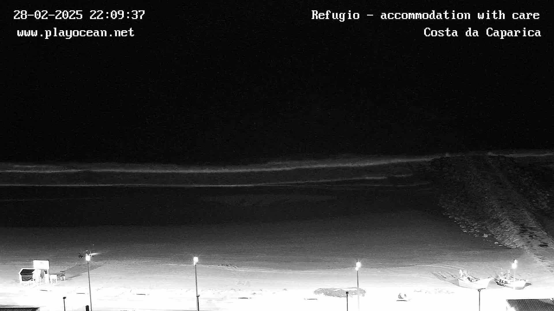 A nighttime beach scene shows dark water with small waves gently lapping the shore,  illuminated by several lights on the beach, with a couple of small boats near a rocky jetty.