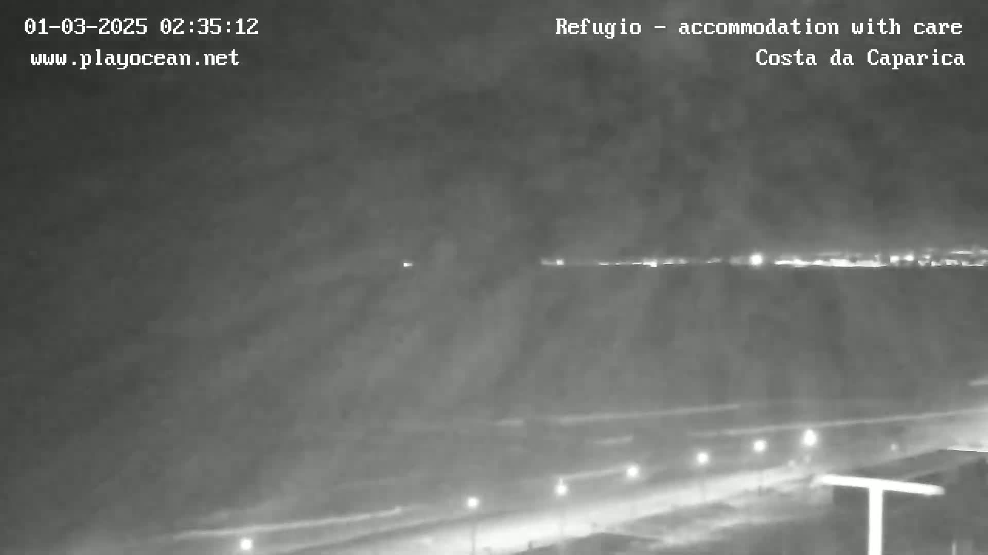 A nighttime, hazy view shows a dimly lit coastal road and distant lights across the water.