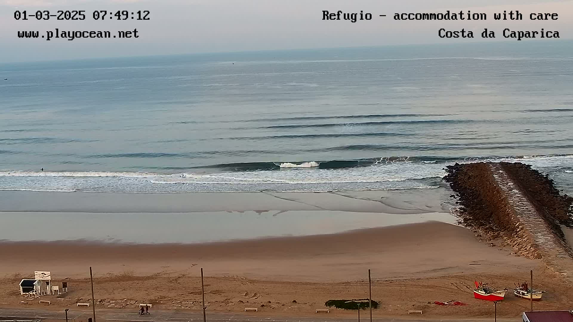 A sandy beach with small waves rolling onto the shore under an overcast sky, featuring a rock jetty extending into the ocean and a few small boats pulled ashore.