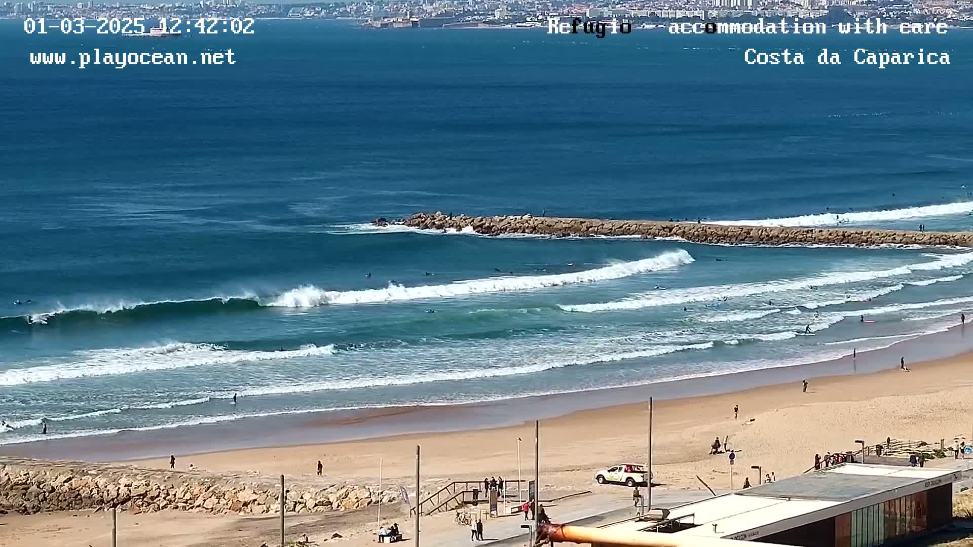 A sunny day at a beach with surfers riding waves that break near a rock jetty, with a city visible in the distance.