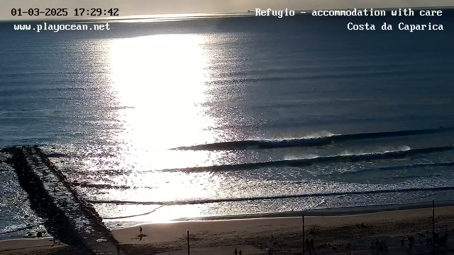 A bright, sunlit ocean with several rolling waves approaching a sandy beach, a jetty, and a few people visible.
