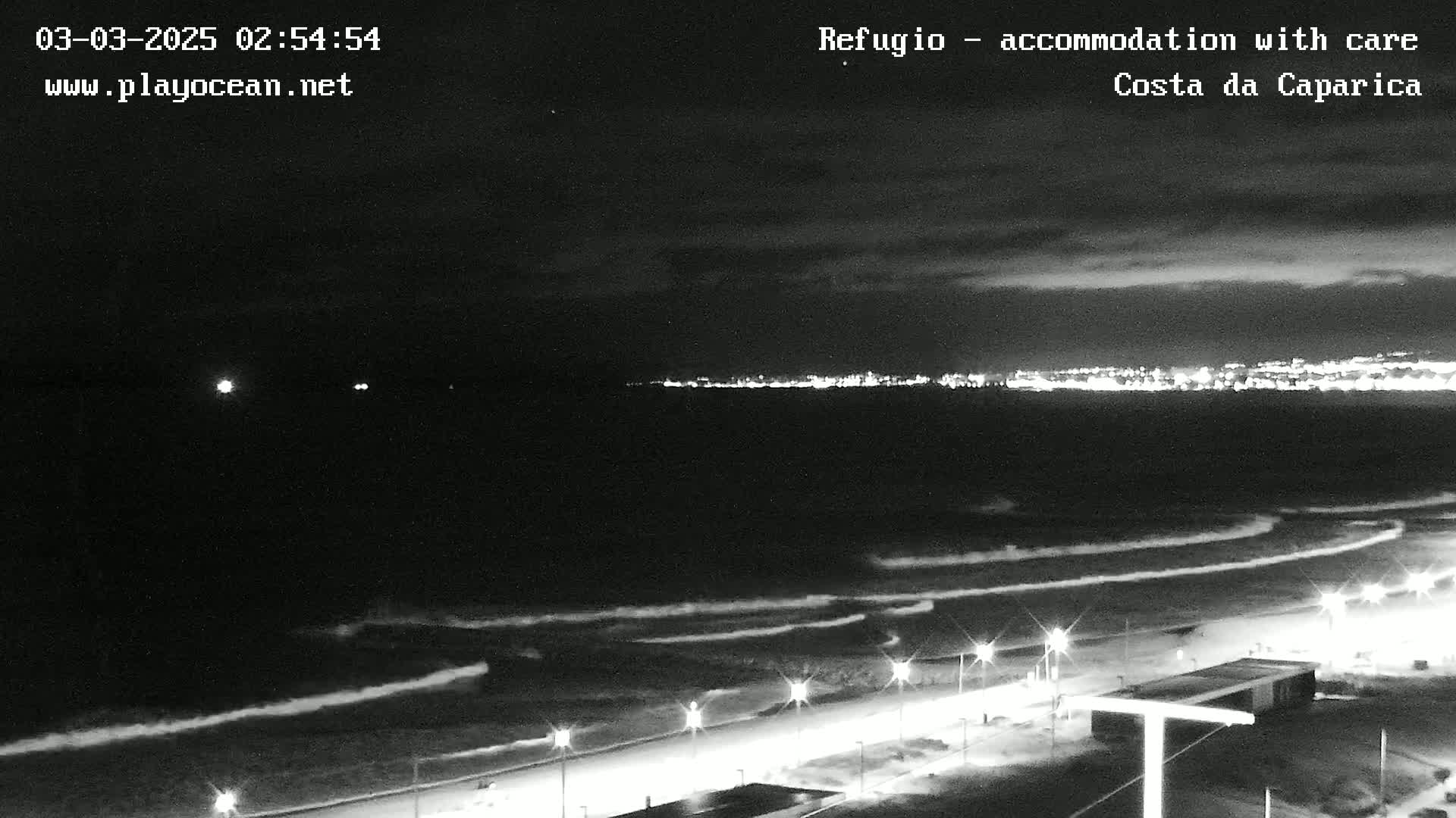 A nighttime beach scene shows a dark ocean with waves lapping the shore under a mostly cloudy sky, with a brightly lit city visible in the distance.