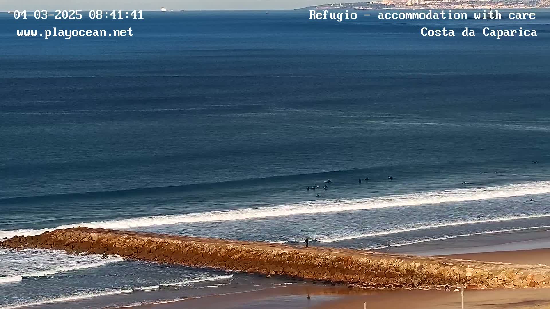 A long rock jetty extends into a blue ocean with several surfers riding small waves on a sunny day.