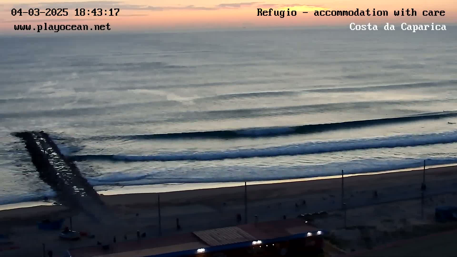 A long, sandy beach at sunset or sunrise, with consistent waves rolling in toward a dark-colored jetty.