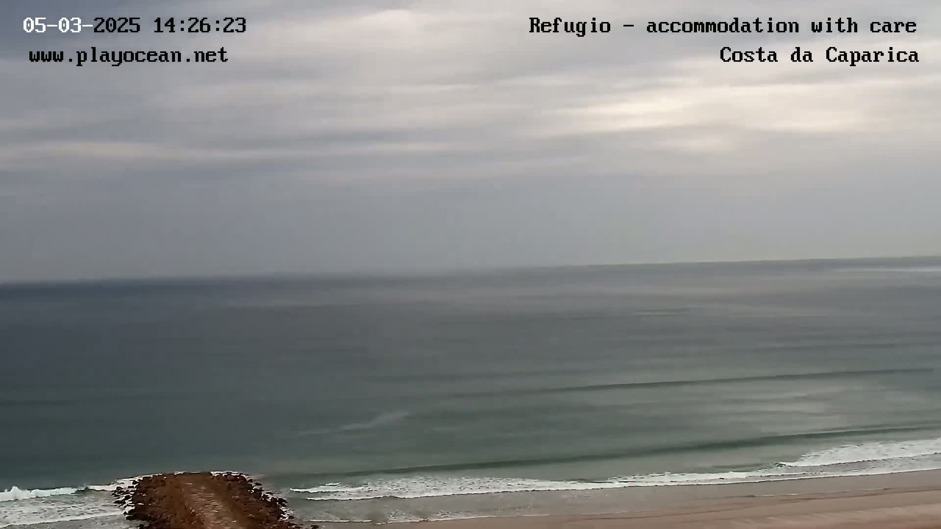 A rocky jetty extends into a gray, overcast ocean with small waves lapping a sandy beach.