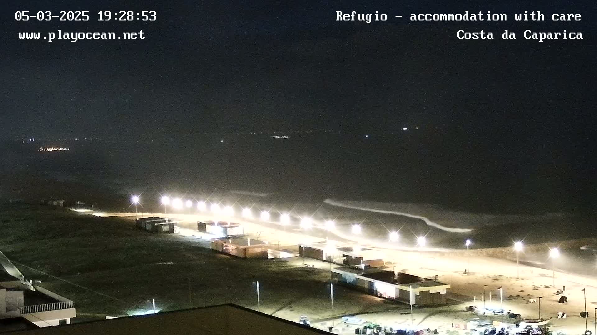 A nighttime view of a beach with gentle waves, illuminated by lights along the shore.