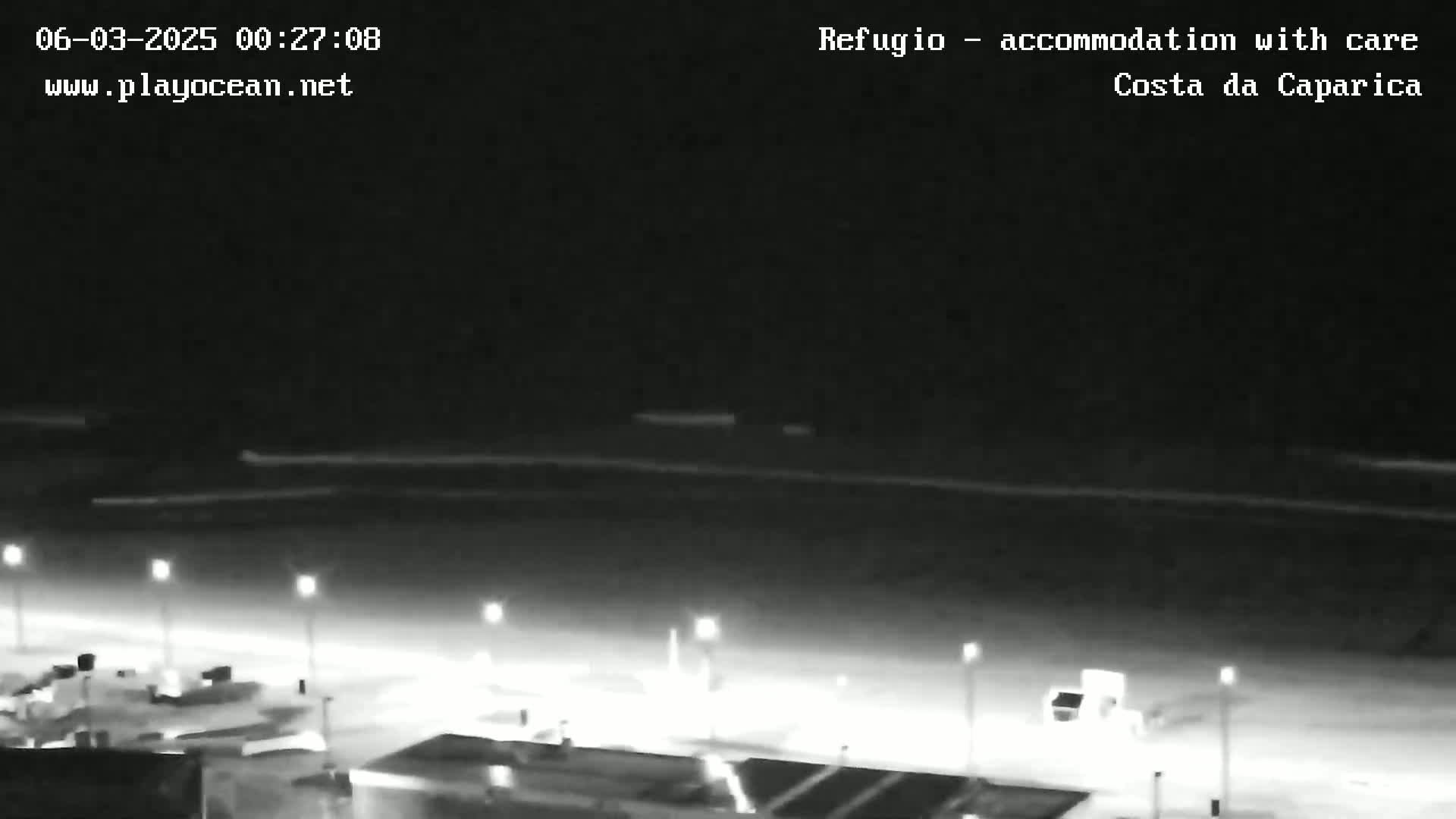 A nighttime view of a dark beach with dimly lit structures and a calm ocean under a clear sky.