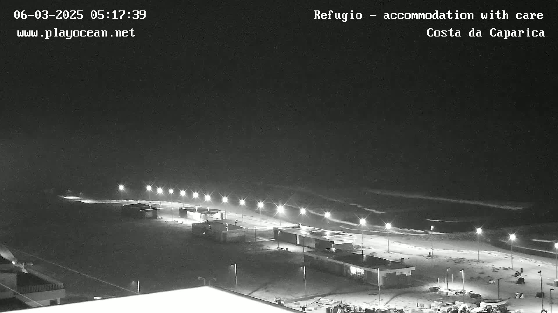 A nighttime, black and white view of a beach at night with gently rolling waves, illuminated by a line of lights along the shoreline.