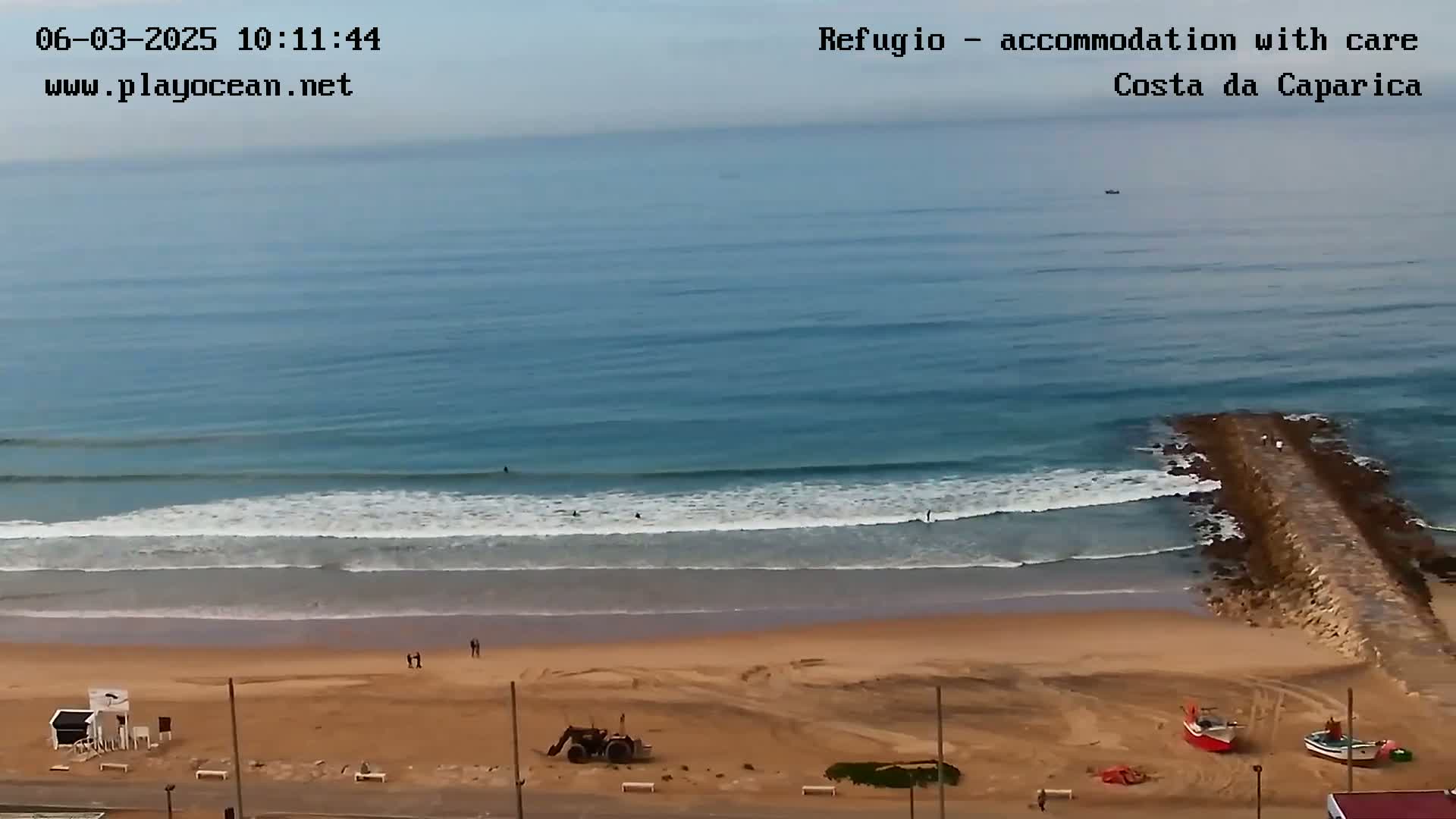 A sandy beach with a few people, a tractor, and small boats, meets a calm ocean with small waves under a mostly clear sky.