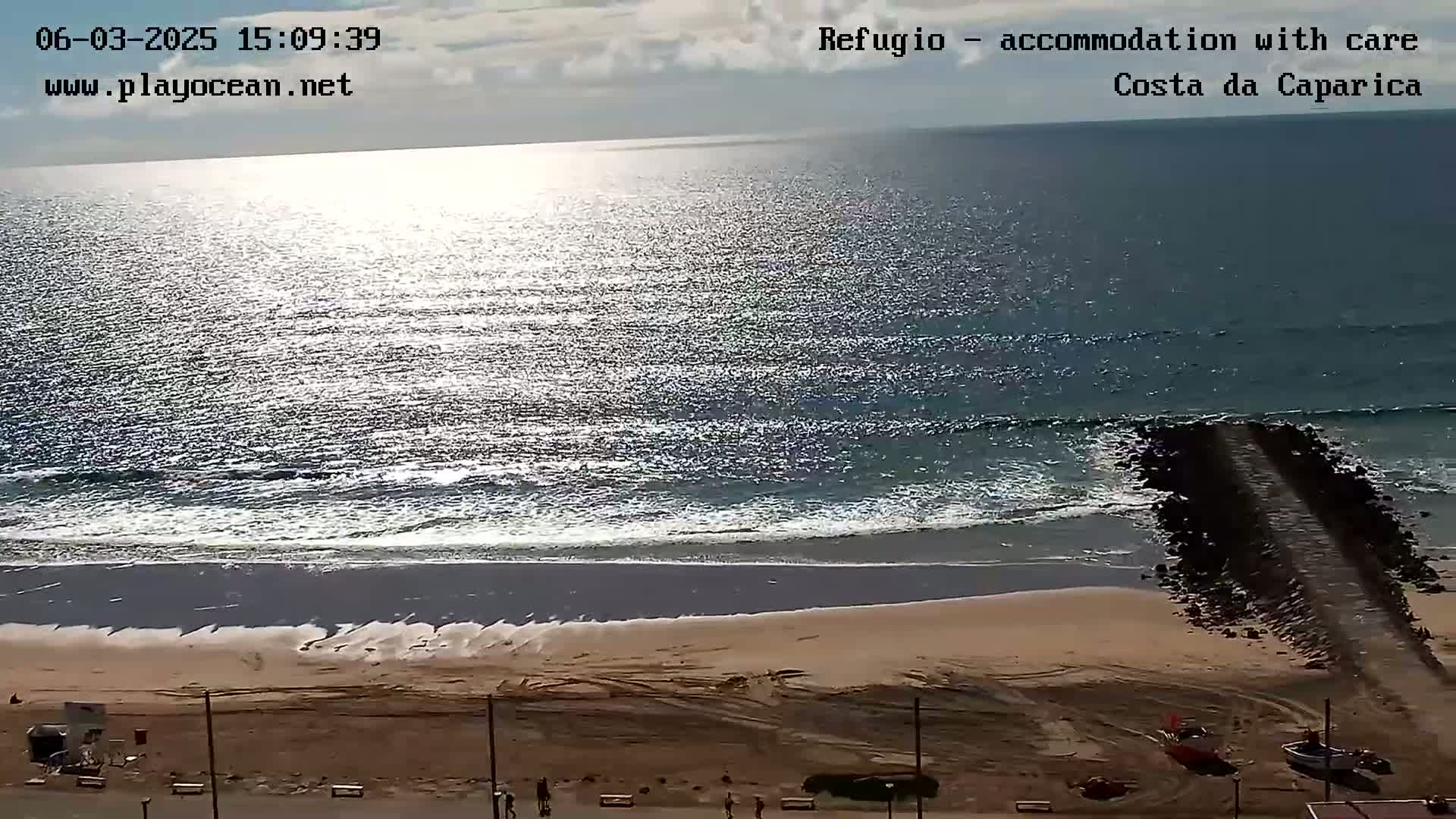A sandy beach with a rocky jetty extends into a calm, sunlit ocean under a partly cloudy sky.