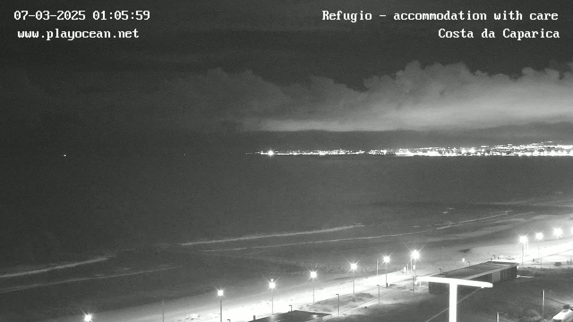 A nighttime coastal scene shows a dark beach with lights along a paved walkway, gentle waves in the ocean, and a distant city skyline illuminated under a partly cloudy sky.
