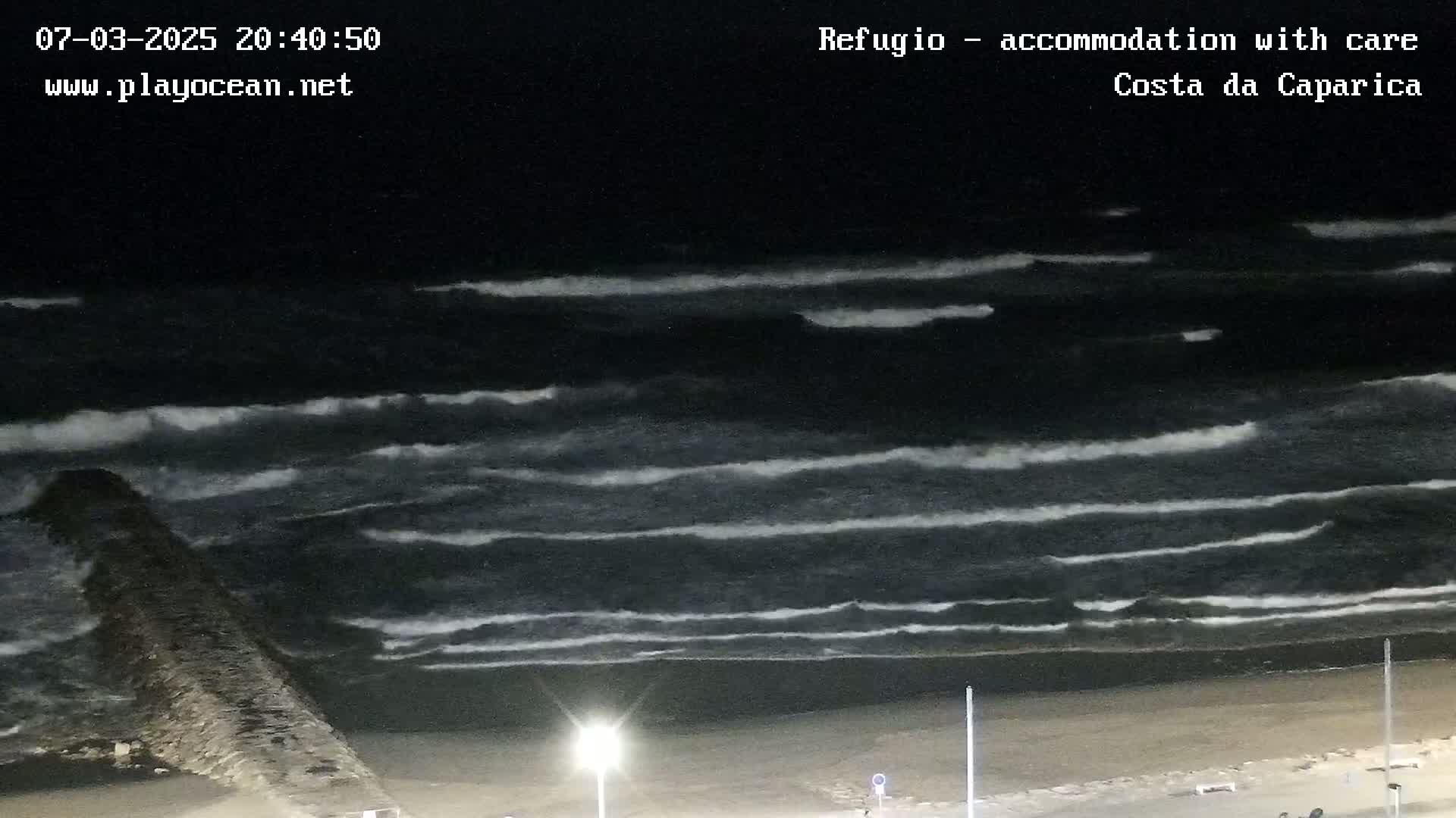 A dark beach at night shows waves rolling onto the shore near a breakwater under a clear, dark sky.