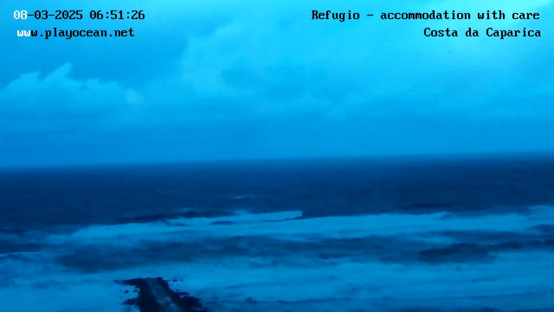 A mostly cloudy, blue-toned sky overlooks a dark blue ocean with moderate waves crashing near a shoreline.