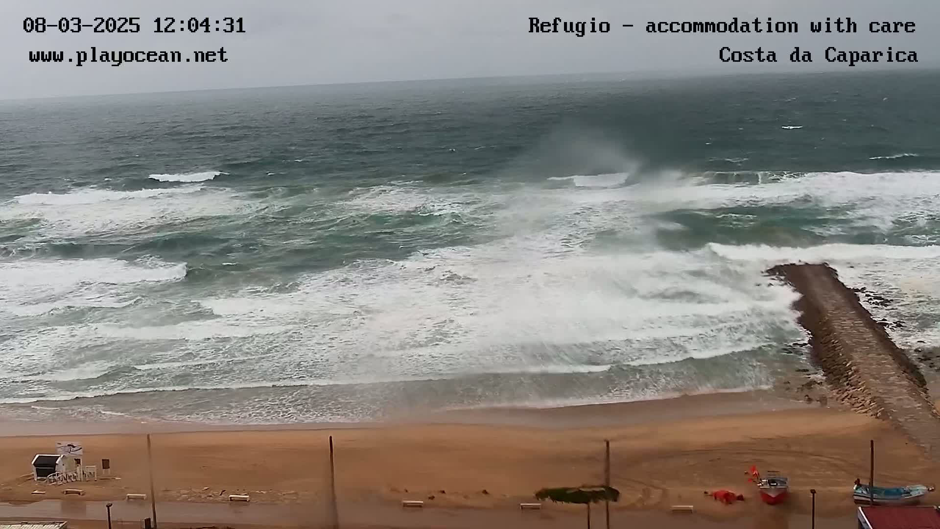 Large waves crash against a beach and a breakwater on an overcast day.