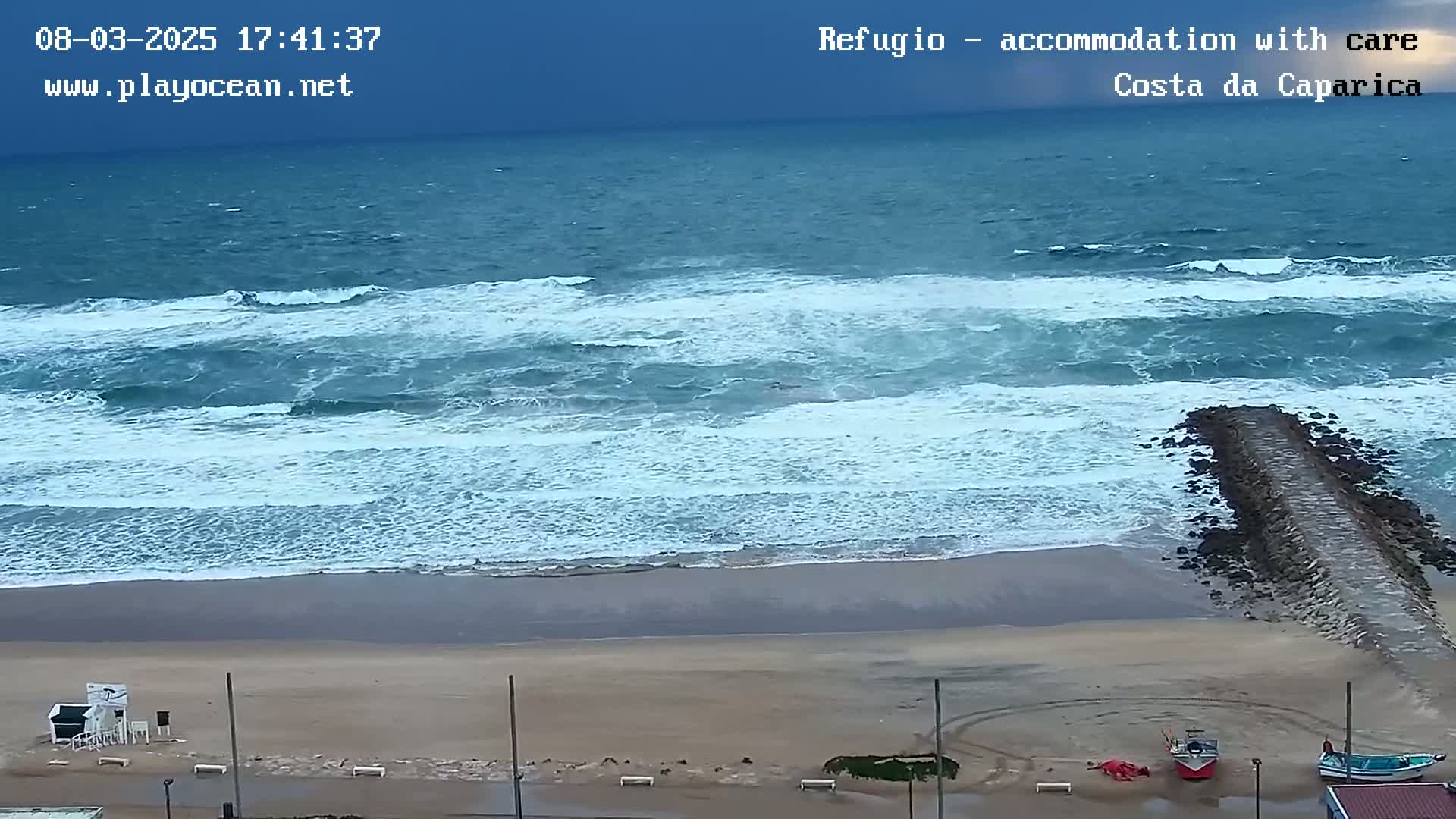 A sandy beach with choppy waves under an overcast sky, a rock jetty extending into the water, and a few small boats pulled ashore.