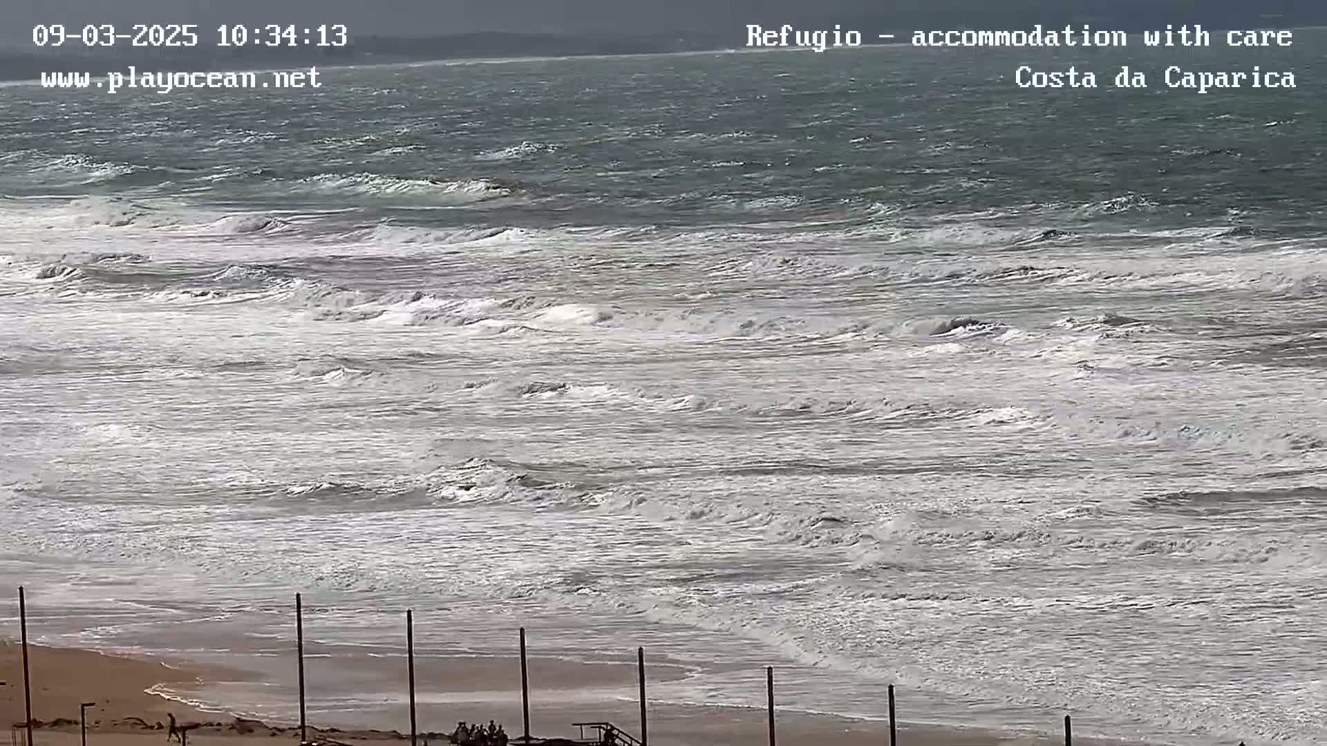 A windy, overcast day shows choppy waves rolling onto a sandy beach with a few people visible near some posts in the distance.