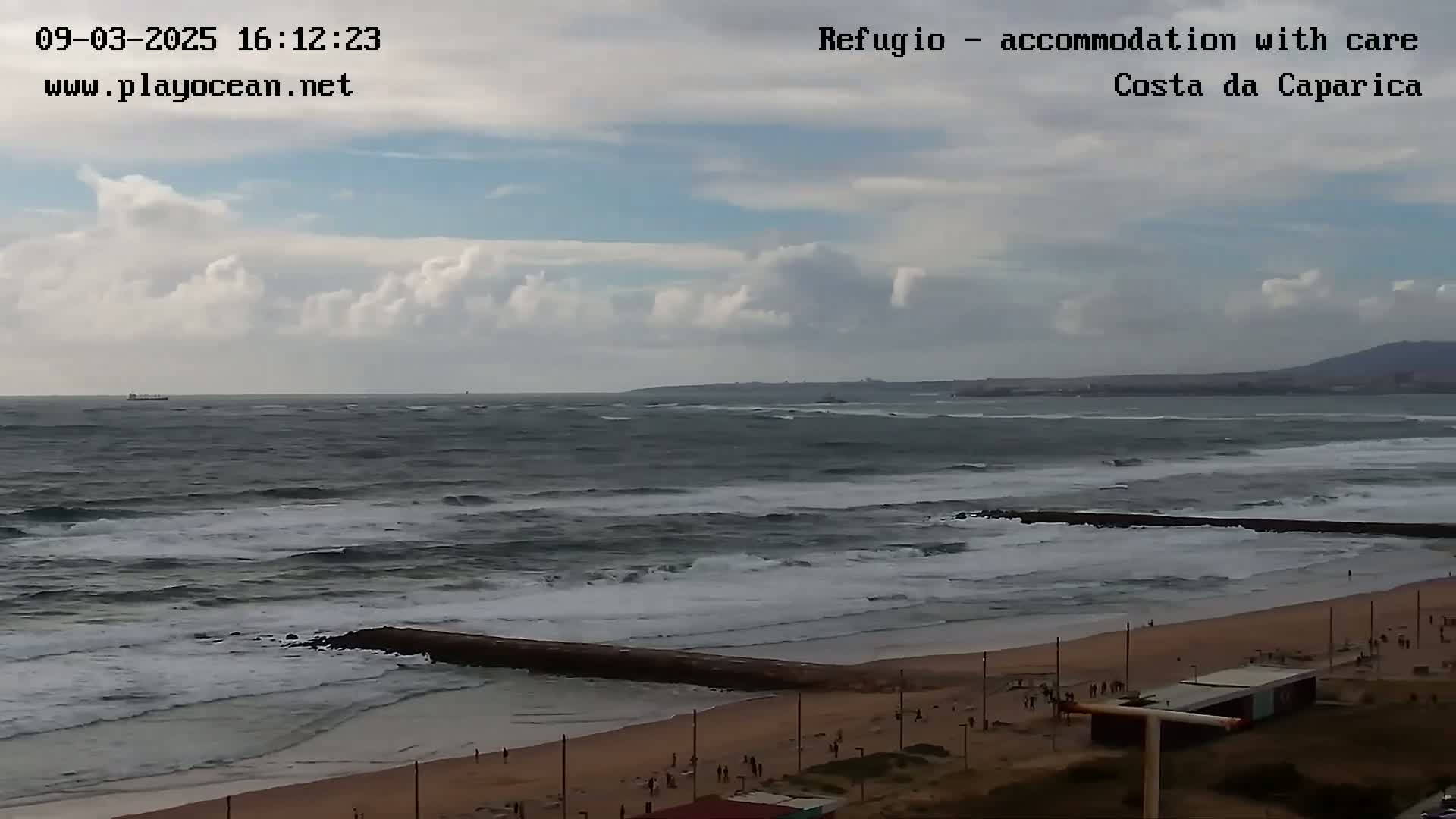 A partly cloudy day shows a sandy beach with a few people, a wave breakwater, and a choppy ocean with some small waves.