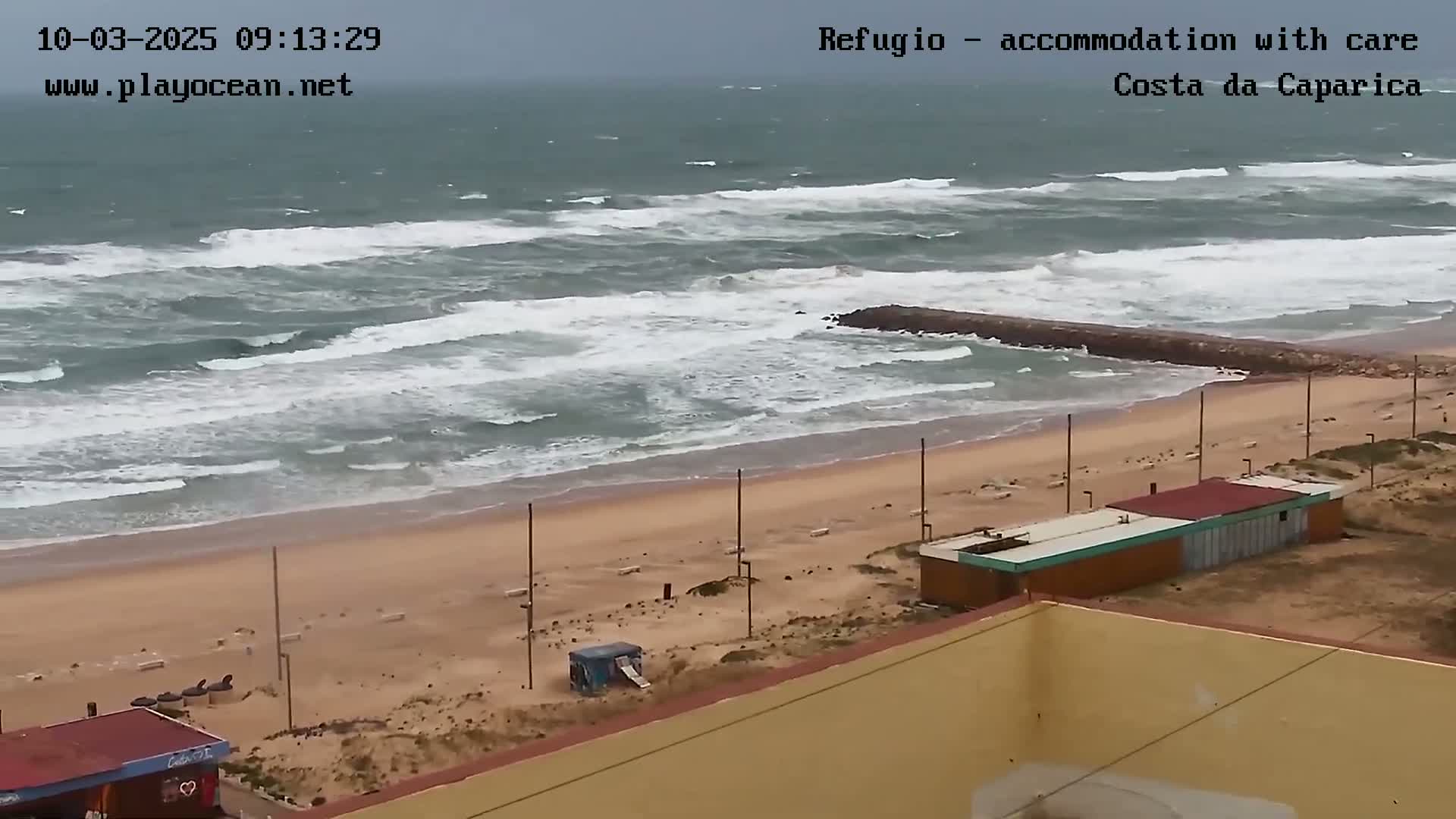 A sandy beach with a few small buildings is shown, with choppy waves and overcast skies in the background.