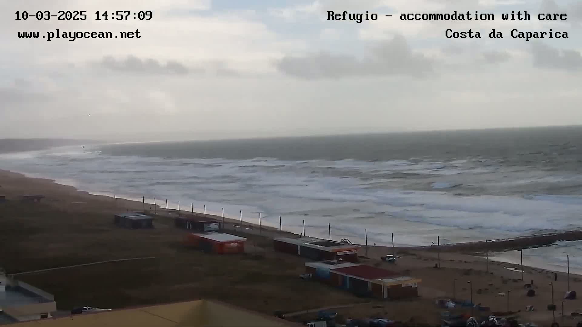 A wide shot of a beach with rough, wavy seas under an overcast sky shows several small buildings near the water's edge.