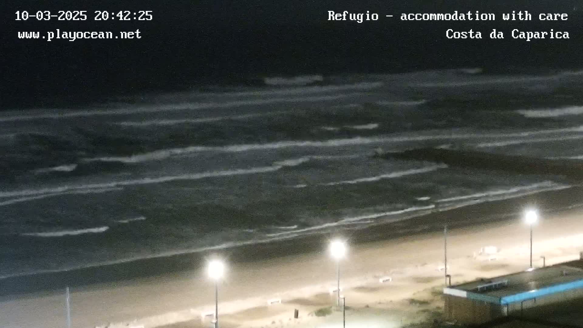 A nighttime beach scene shows dark, moderately wavy ocean water lapping a sandy shore illuminated by streetlights.