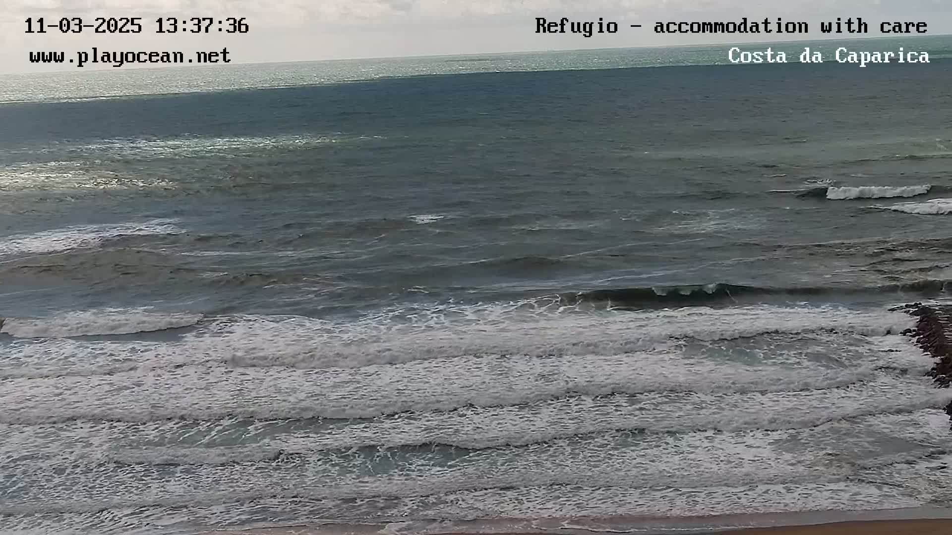 A mostly cloudy day shows dark-grey ocean waves breaking on a sandy shore.