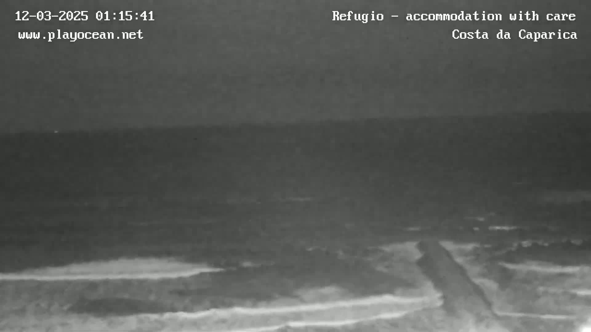 A nighttime, low-light view of a dark ocean meeting a beach with a dark, possibly paved path visible in the foreground.