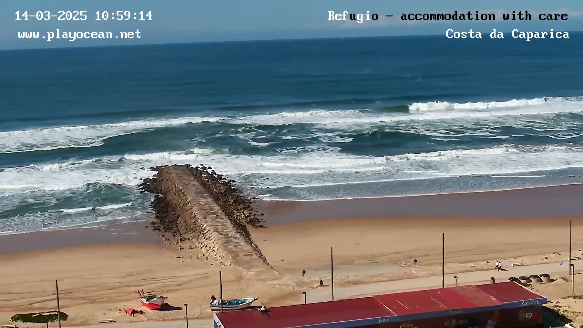 A sandy beach with a long rock jetty extending into a wavy ocean under a clear, sunny sky.